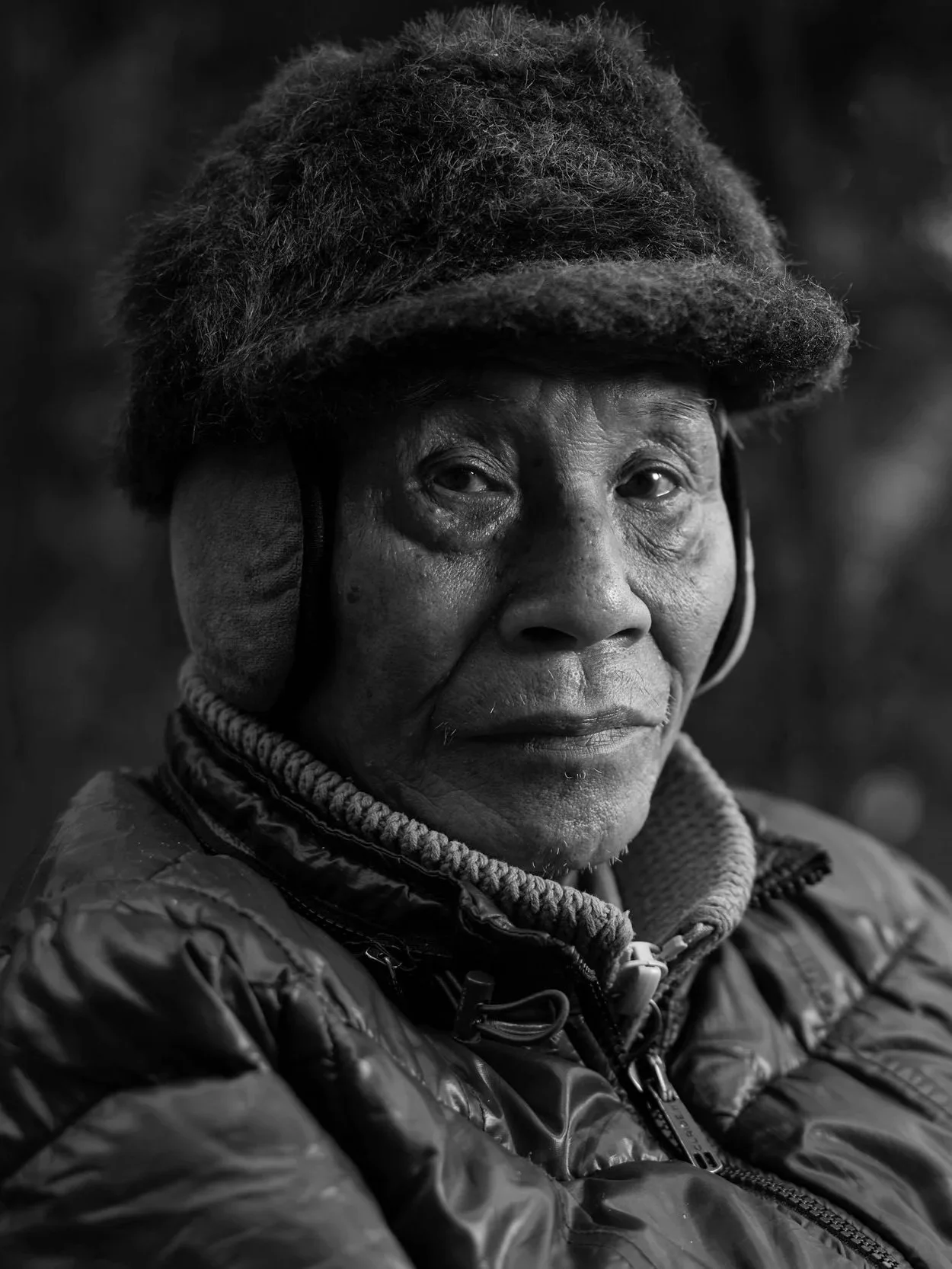 Black and white photo of an elderly woman wearing a hat and a quilted jacket, looking slightly to the side with a serious expression, outdoors with blurred trees in the background.