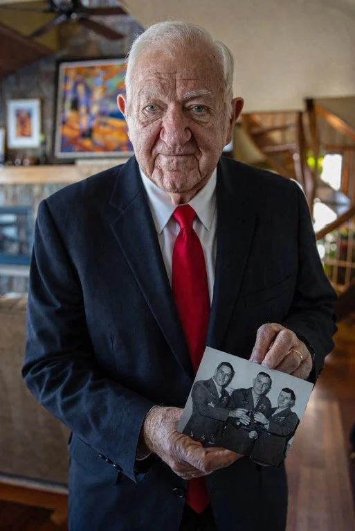 An elderly man with white hair, wearing a black jacket over a red shirt, sitting in a room with bookshelves in the background, holding a framed black-and-white photo of three men in uniform.
