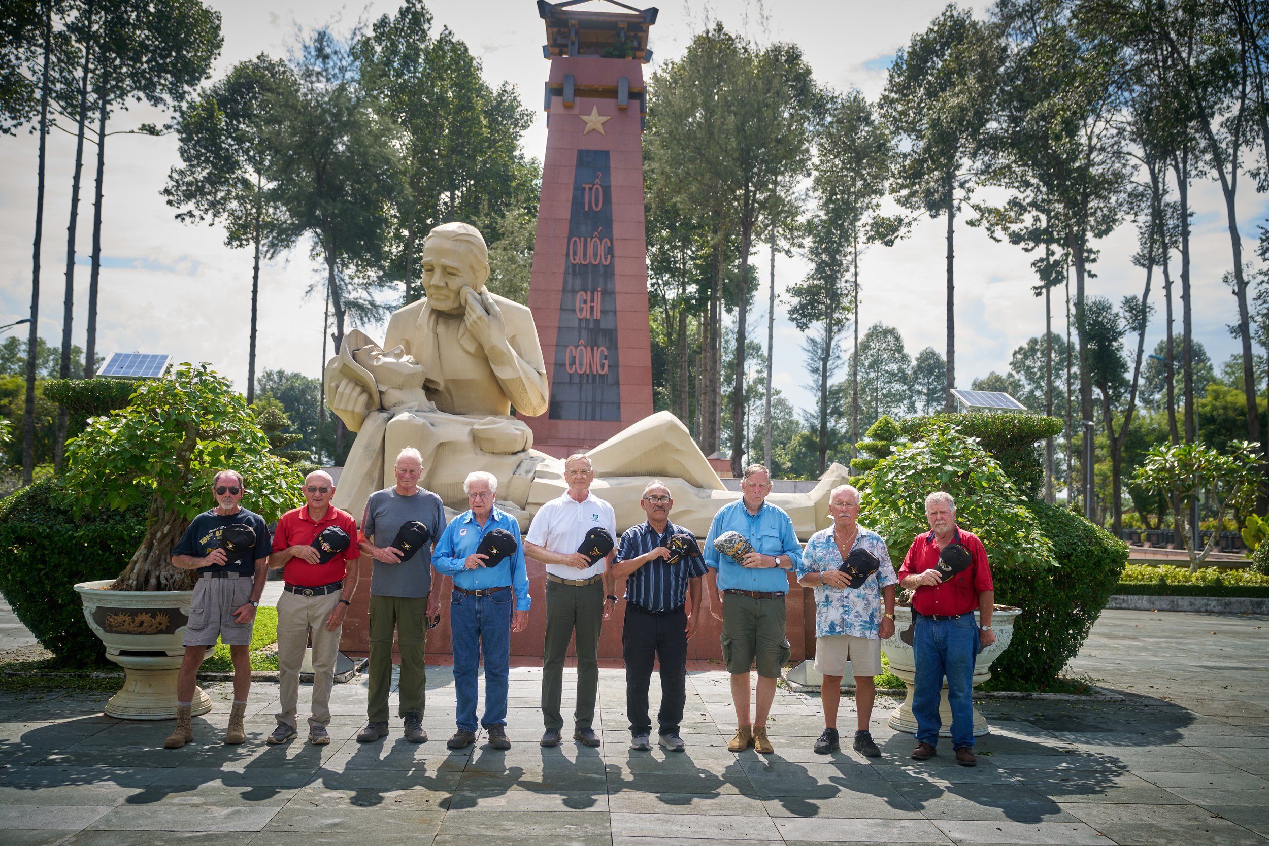 A group of eleven men holding hats stands in front of a large statue and monument with trees in the background."}