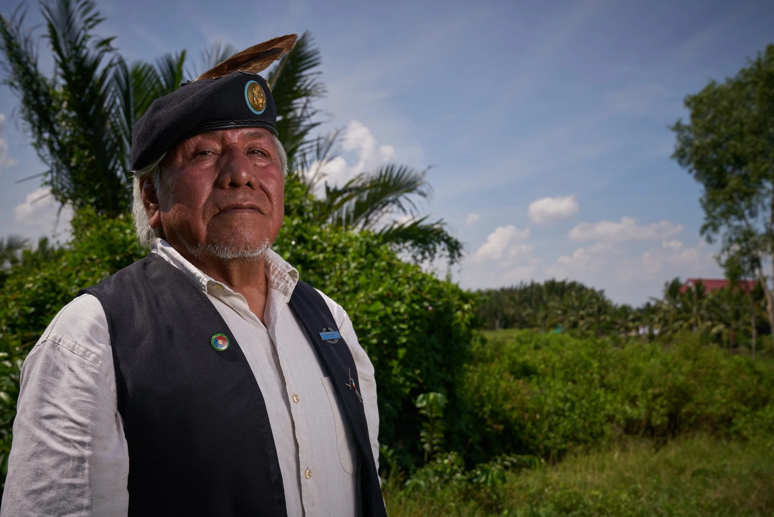An elderly indigenous man wearing a black feathered headdress, white collared shirt, and black vest stands outdoors amidst greenery, with a blue sky and clouds in the background.
