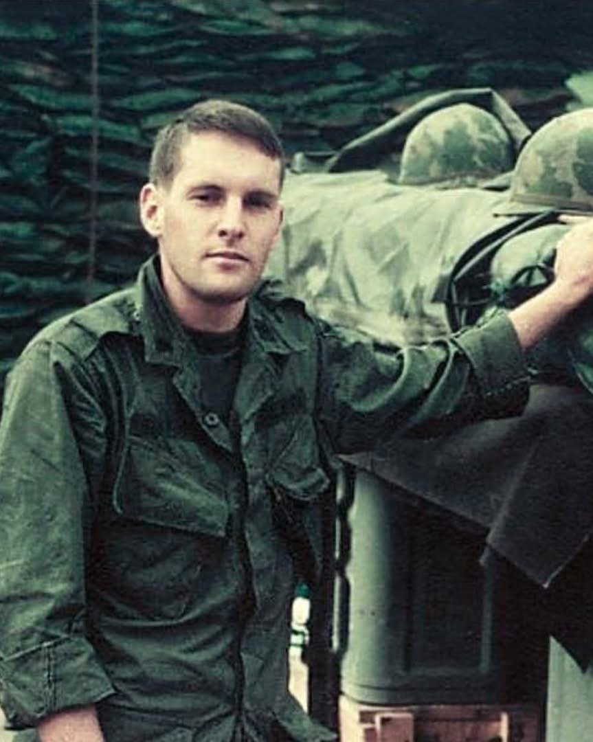A soldier wearing a military uniform stands next to a camouflage netting and military equipment. He has short hair and a neutral expression. The background features sandbags and helmets.