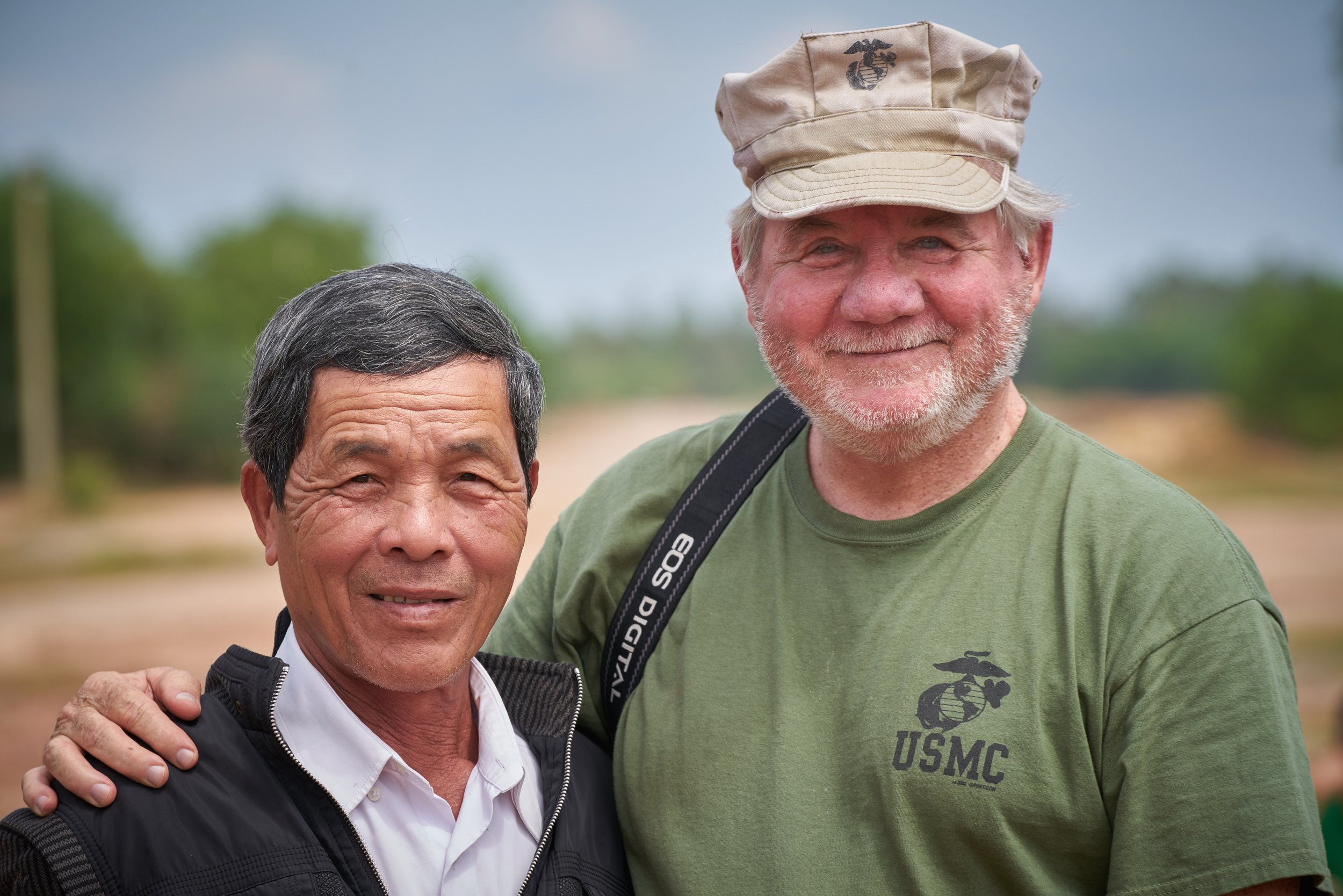 Two men smiling outdoors; one wearing a green "USMC" shirt and beige cap, the other in a black jacket with a white shirt. Background is blurred greenery and sky.