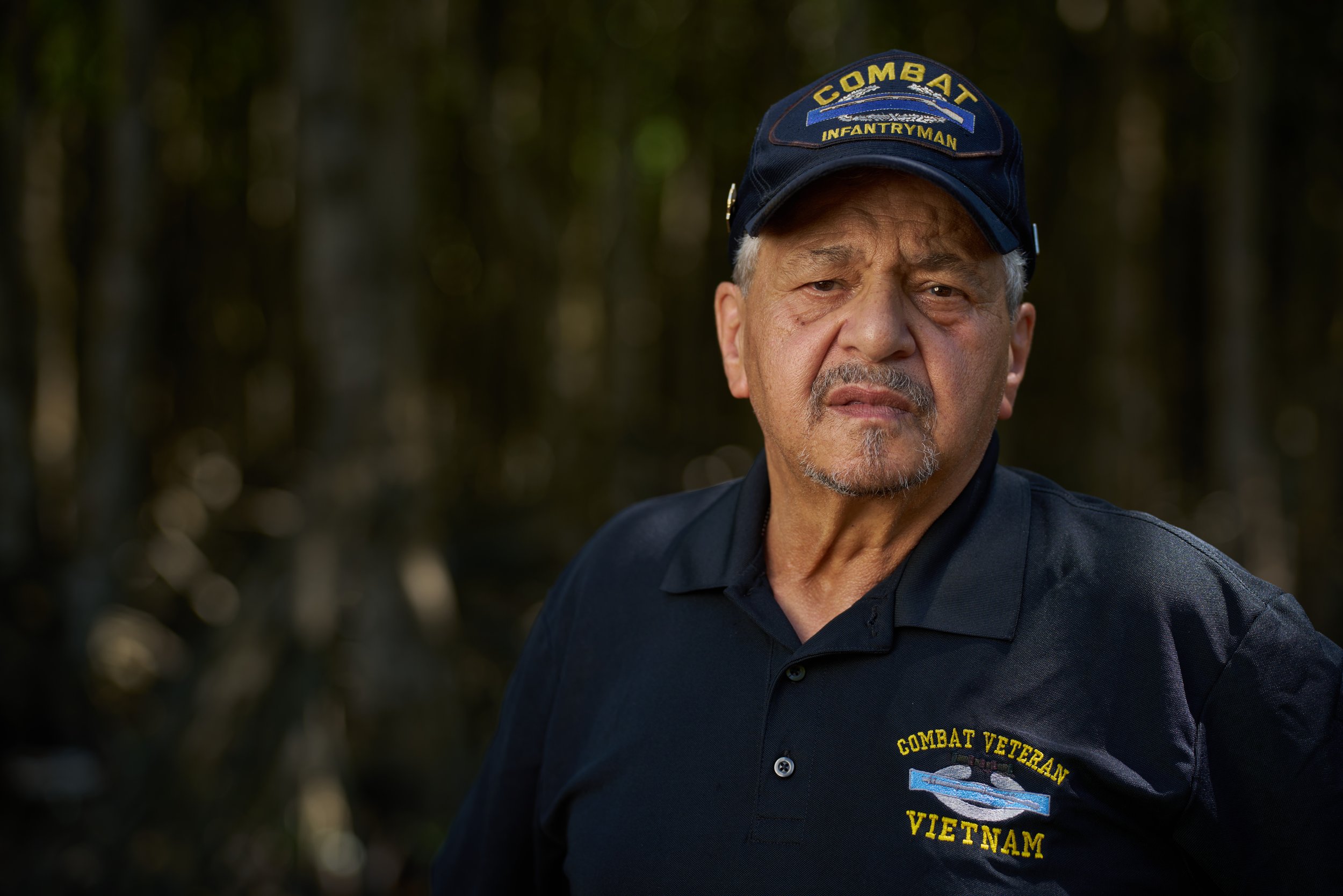 A man wearing a "Combat Infantryman" cap and a "Combat Veteran Vietnam" shirt standing outdoors with a forested background.