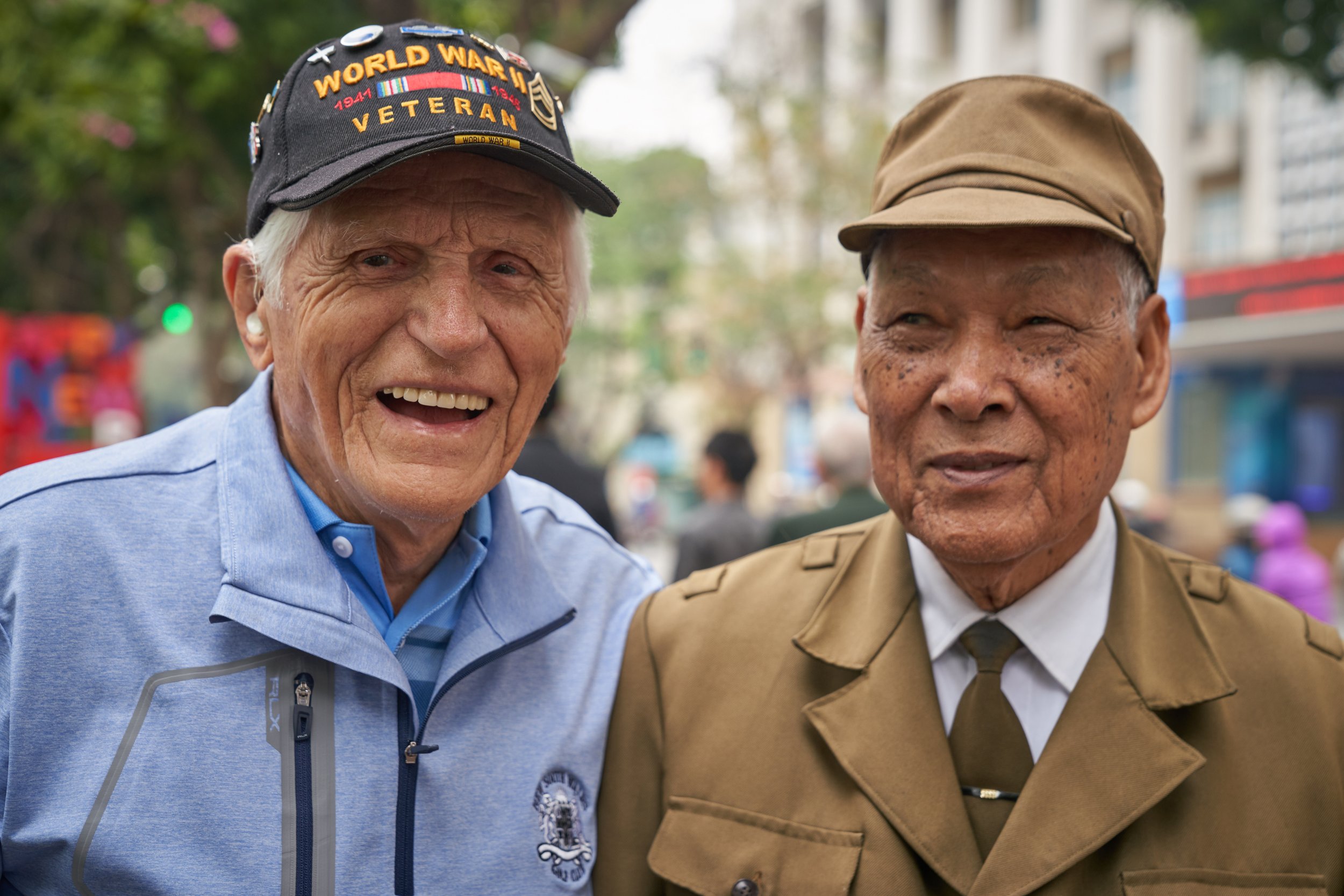 Two elderly men standing together, one wearing a "World War II Veteran" cap and a light blue jacket, the other in a brown military-style uniform and cap, outdoors with a blurred street background.