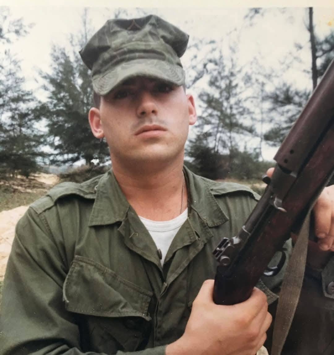 A person in military uniform holding a rifle in an outdoor setting with trees in the background.