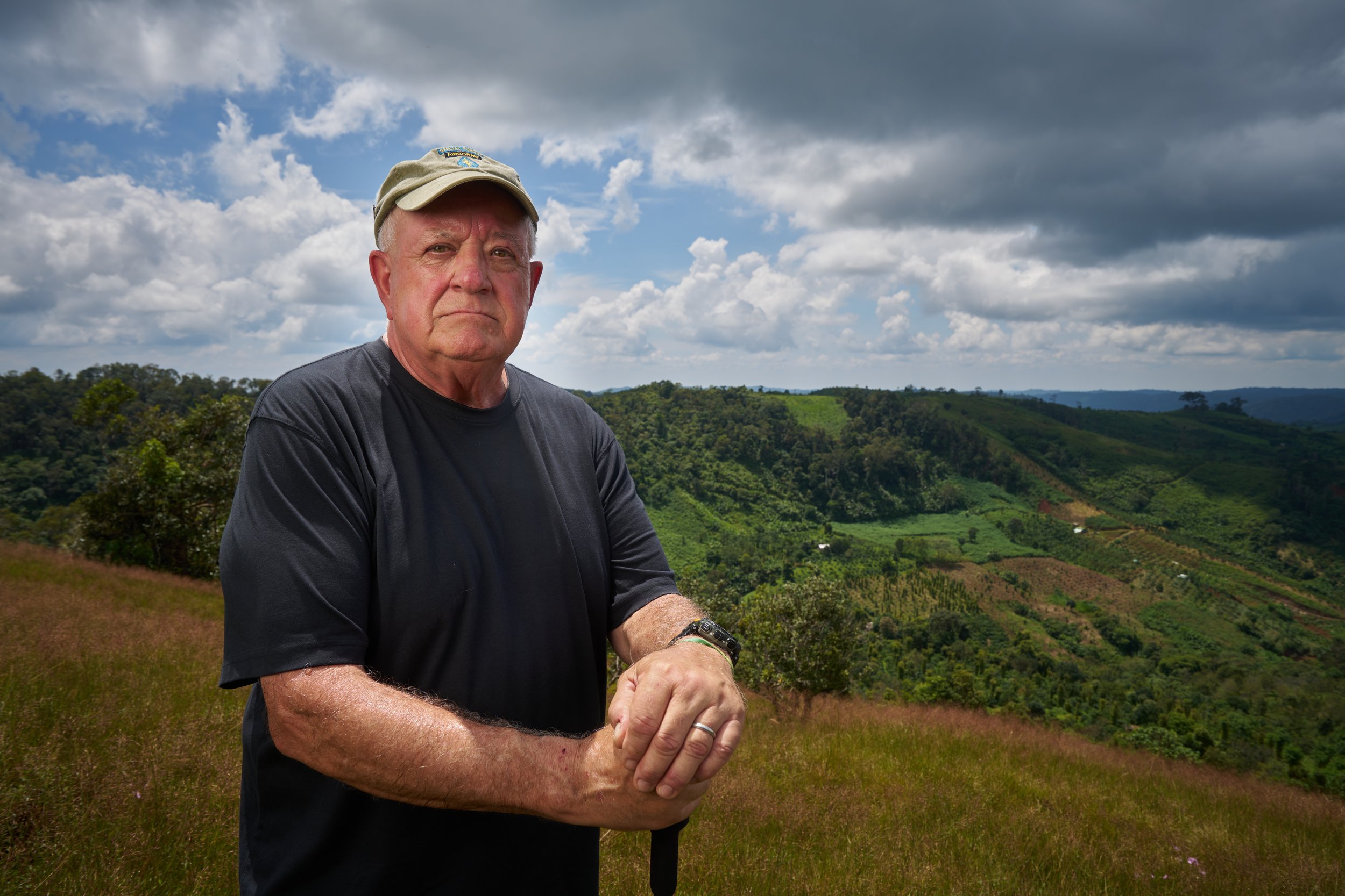 Elderly man wearing a cap and black shirt standing in front of a lush green hill under a cloudy sky.