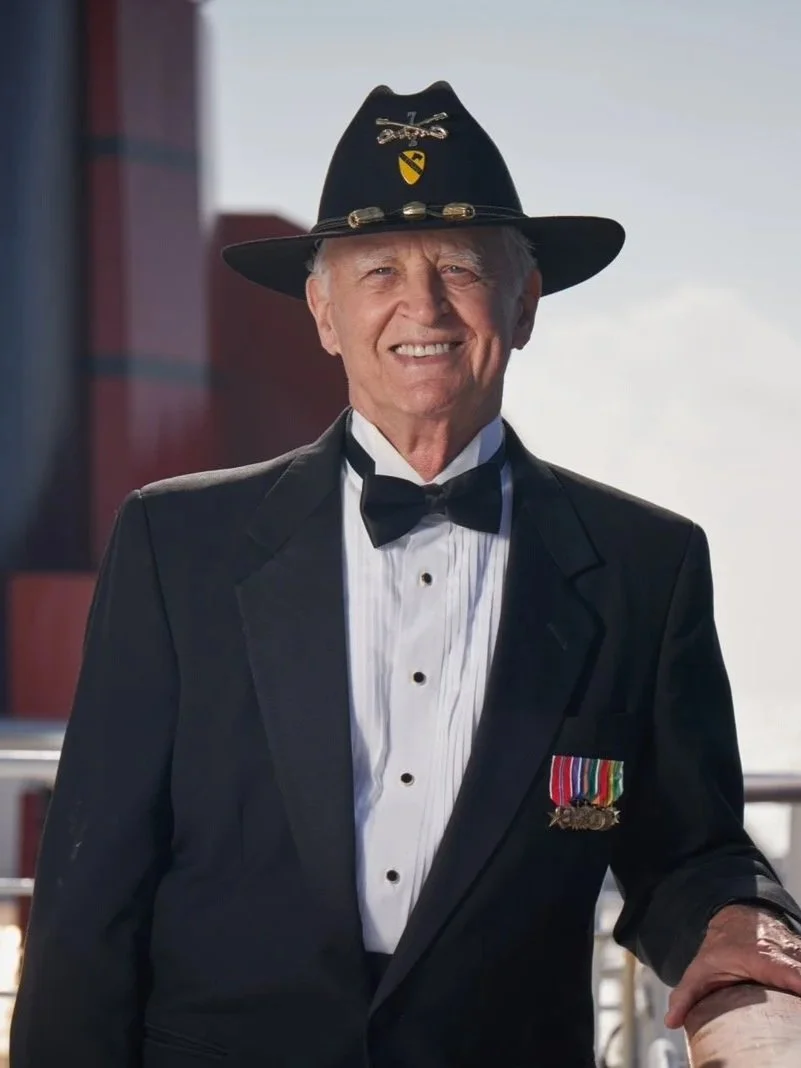 An elderly man dressed in formal black tuxedo with a bow tie, wearing military medals on his chest, sitting indoors against a dark background.