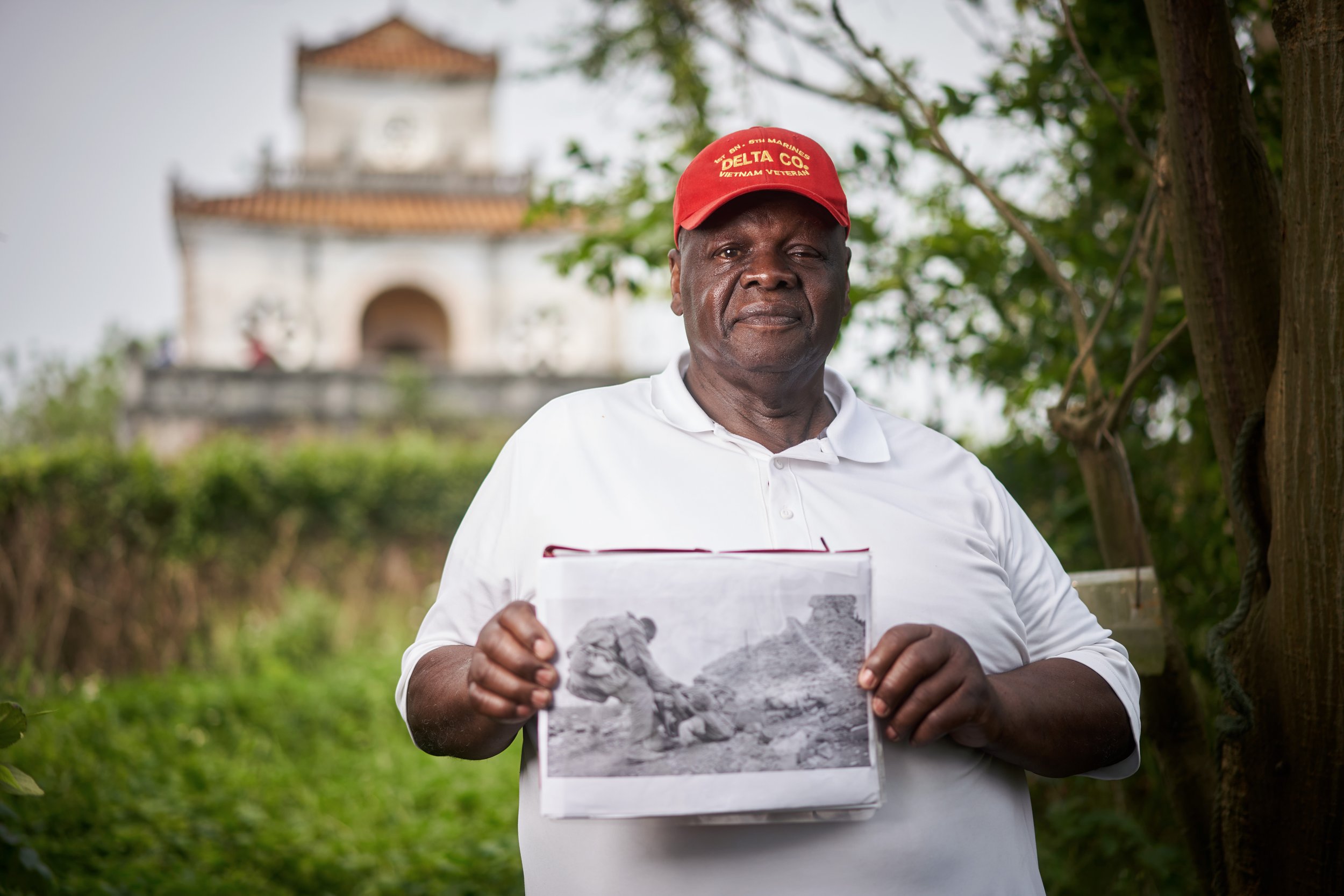 A man wearing a red cap and white polo shirt holds a black and white photo in an outdoor setting with a historical building in the background.