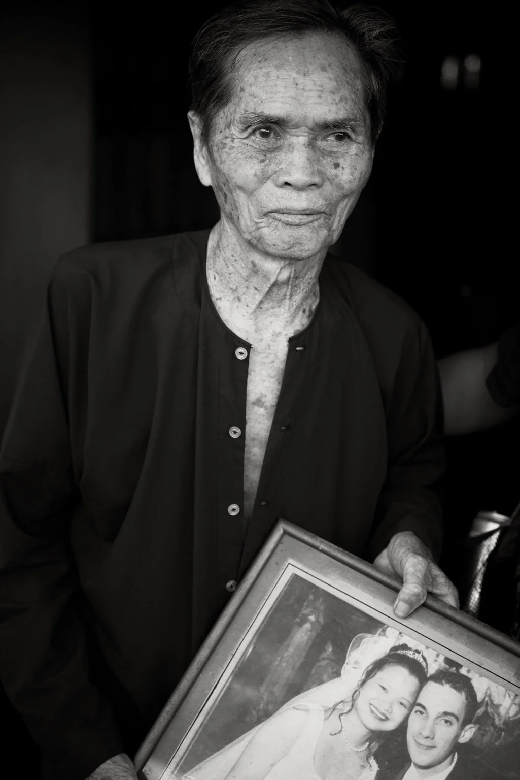An elderly woman with age spots on her face holds a framed photograph of a smiling couple, possibly a wedding photo, in a black and white image.