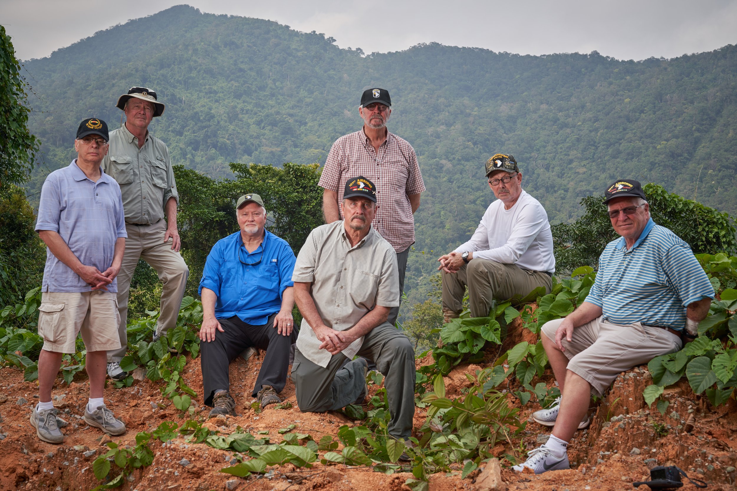 A group of seven men wearing casual outdoor clothing and hats pose in a hilly, green forested area. There is a mountain visible in the background.