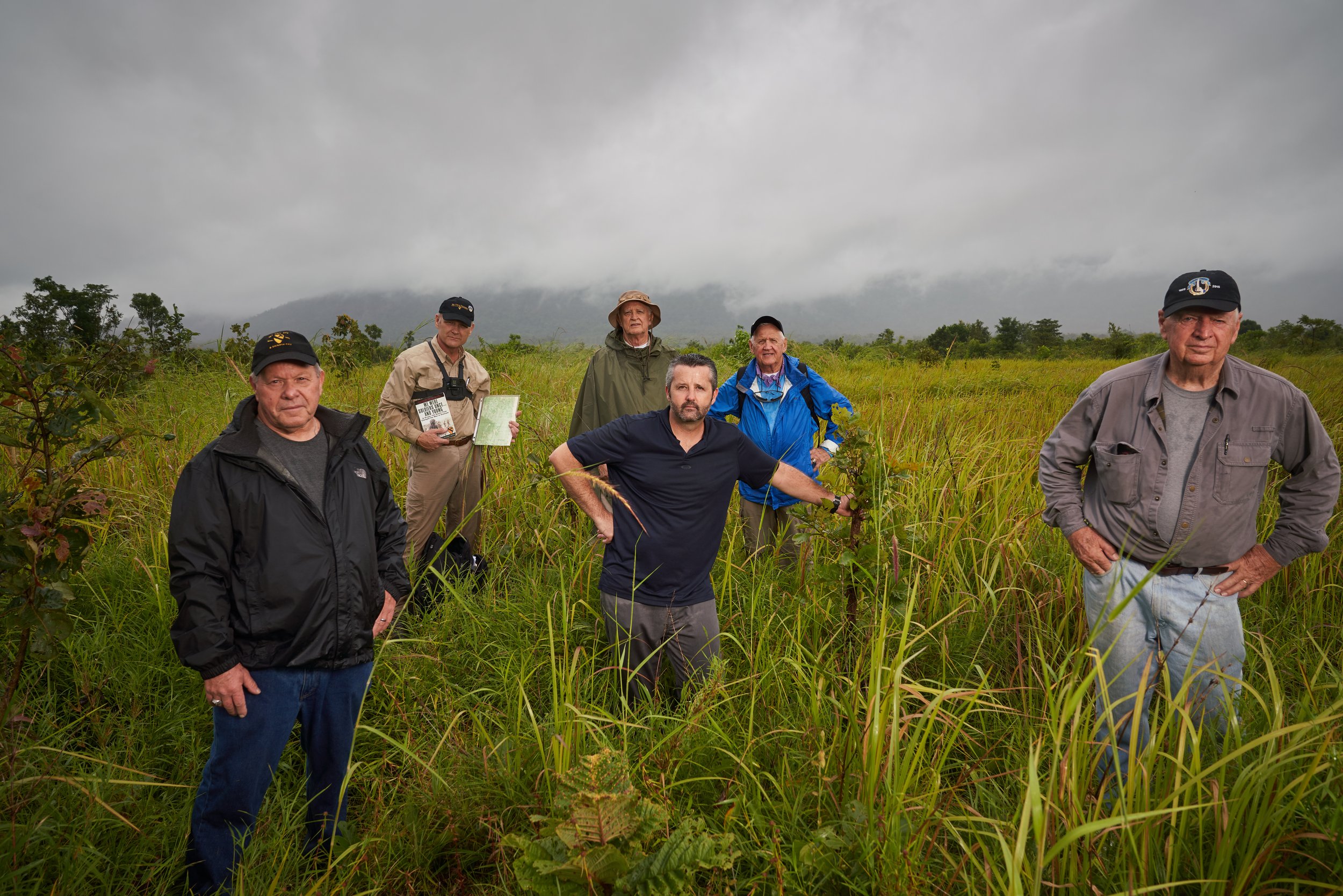 Group of six men standing in a grassy field under cloudy skies.