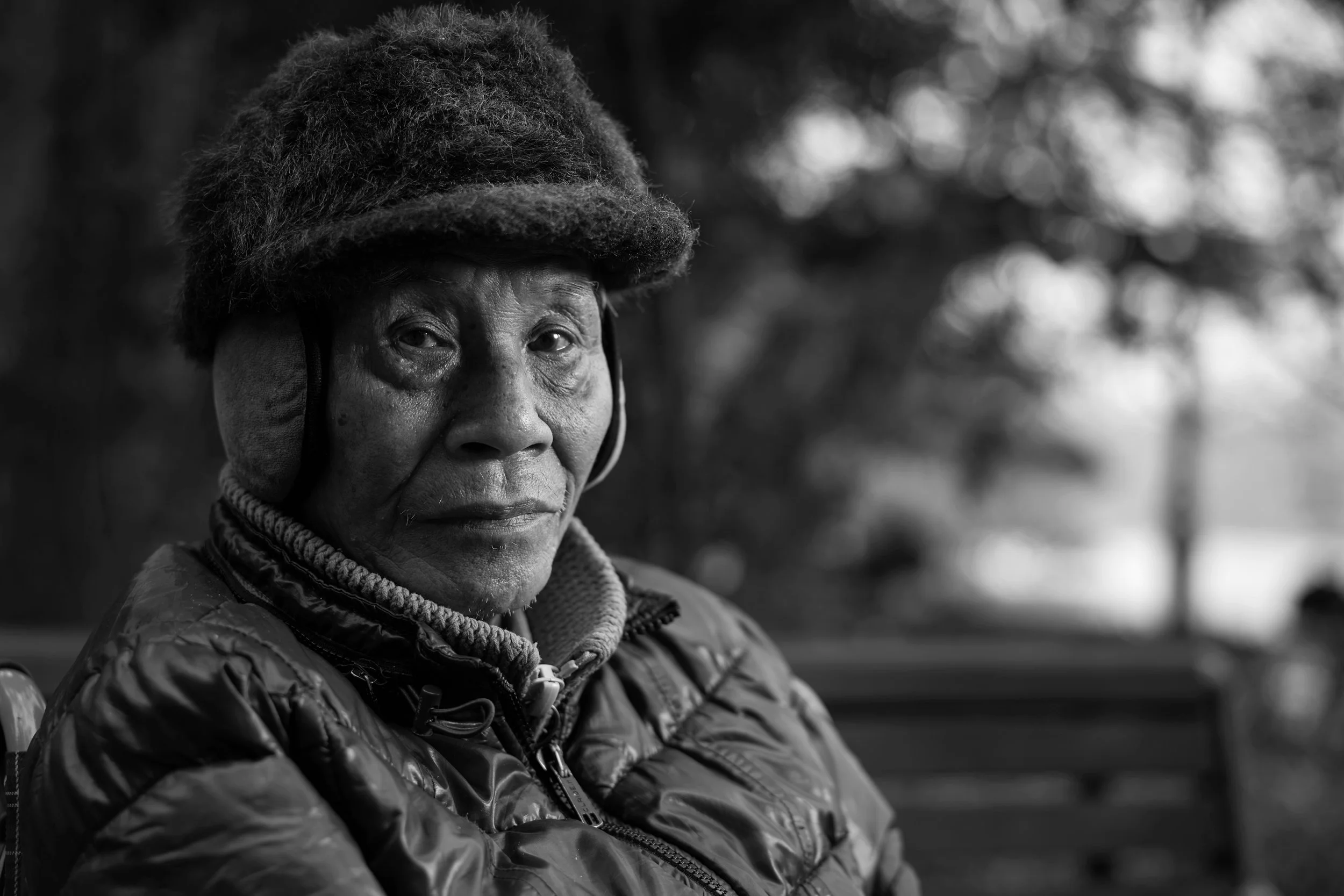 Black and white photo of an elderly woman wearing a hat and a quilted jacket, looking slightly to the side with a serious expression, outdoors with blurred trees in the background.