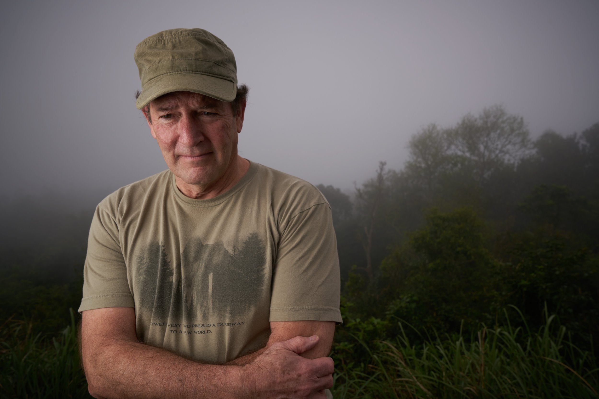 A man wearing a hat and t-shirt stands with arms crossed in a misty outdoor landscape with trees in the background.