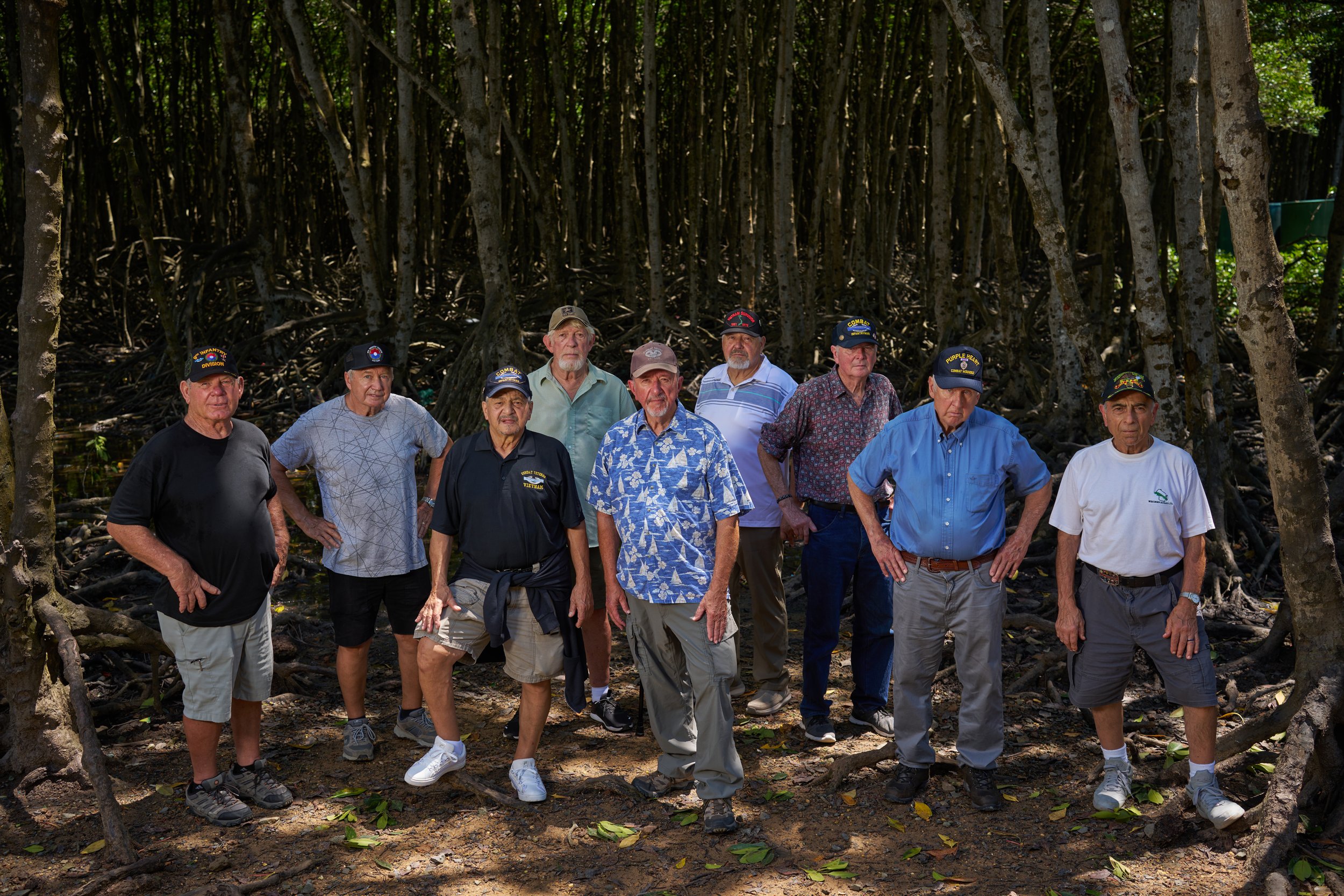 Group of older men outdoors surrounded by trees, wearing casual clothing and hats.