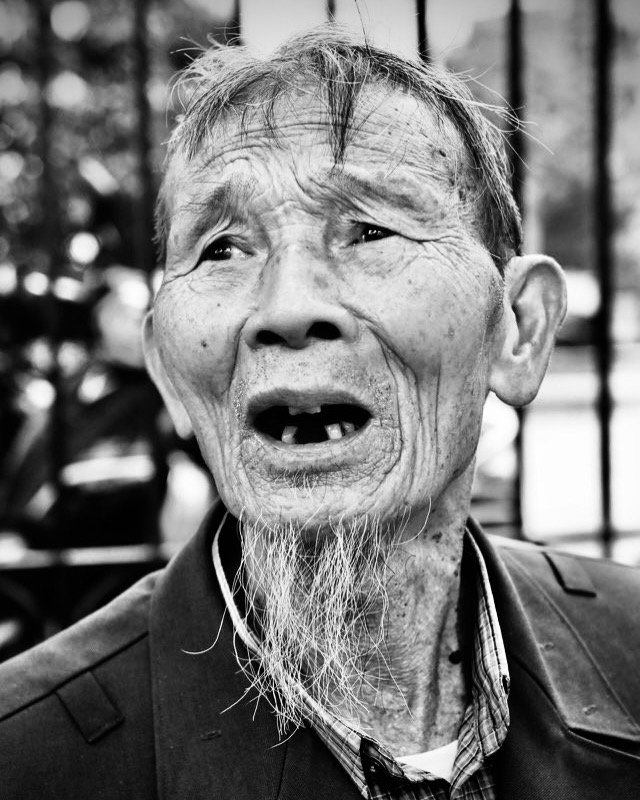 An elderly man with missing teeth, a long beard, and a surprised or expressive facial expression. He is wearing a suit jacket and shirt, with a blurred background.