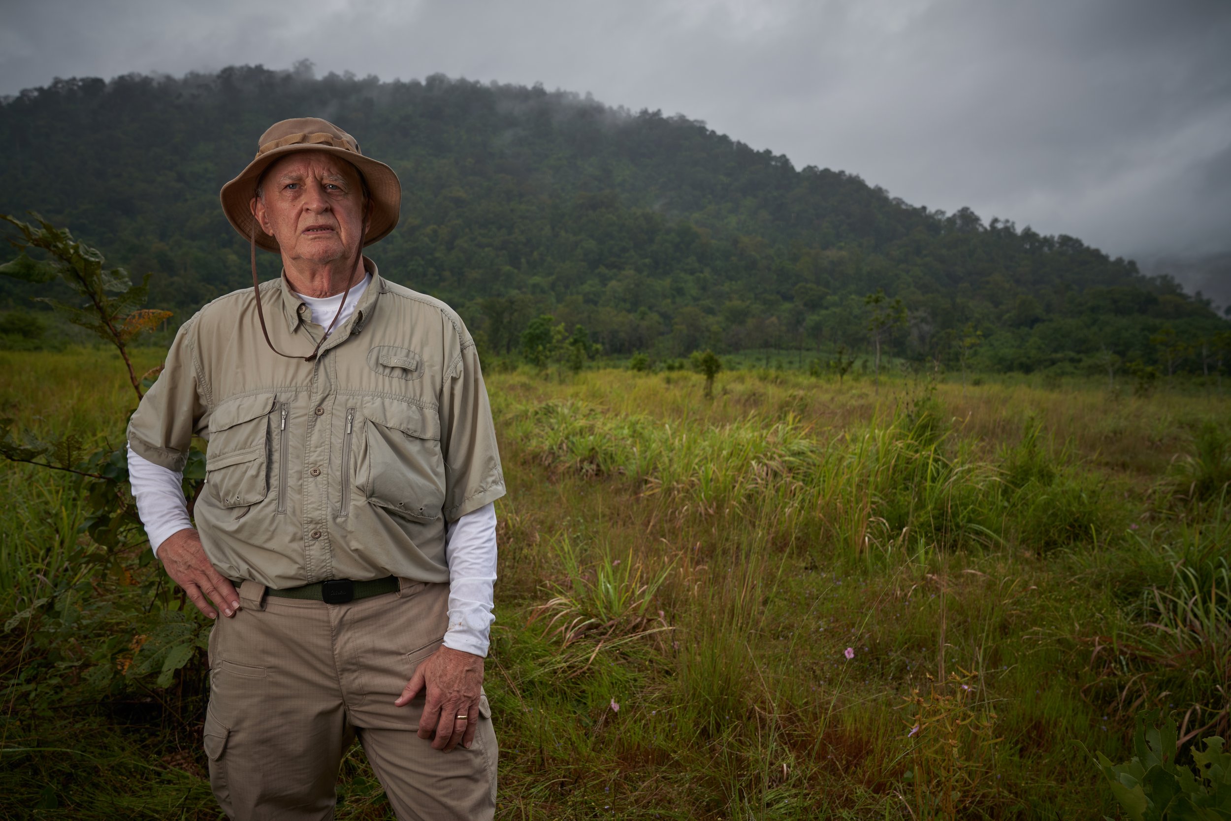 Elderly man in outdoor attire standing in a grassy field with a forested mountain background.