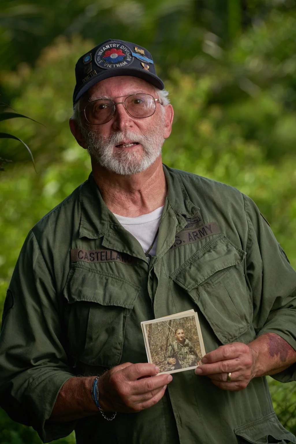 An elderly man with glasses and a white beard, wearing a dark cap with patches and a green military jacket, stands outdoors in a wooded area, holding a photograph of a young soldier in uniform.