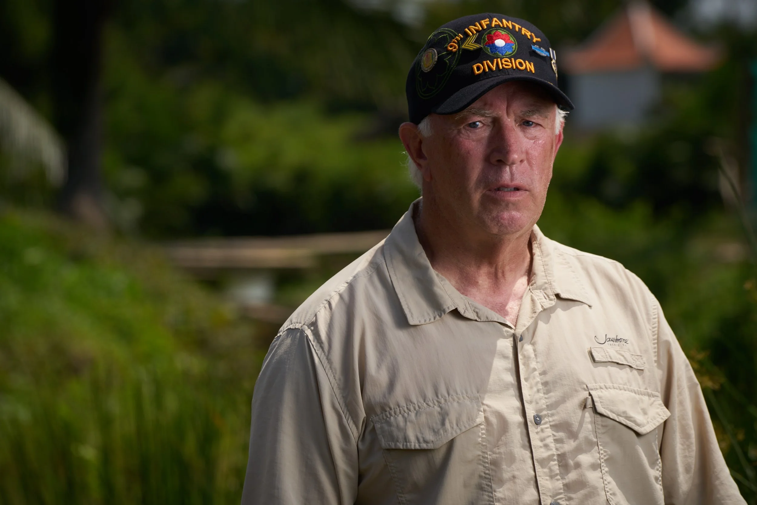 A man wearing a beige button-up shirt and a black cap with patches and the words "Our Infantry Division" visible on it.