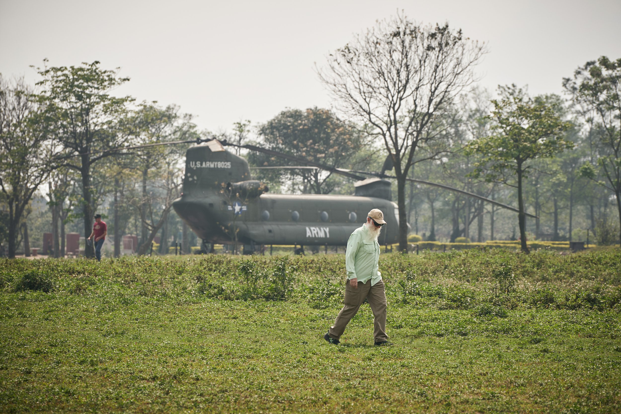 A person walks in a grassy field with a military helicopter, labeled "U.S. Army," in the background. Trees and a few other people are visible in the distance.
