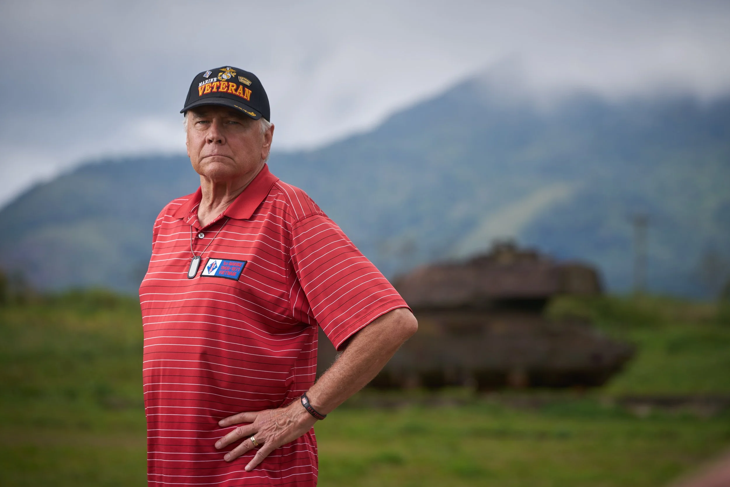 An elderly man wearing a black cap with patriotic insignia, a red polo shirt with white stripes, and a patriotic flag patch, standing outdoors with mountains and cloudy sky in the background.
