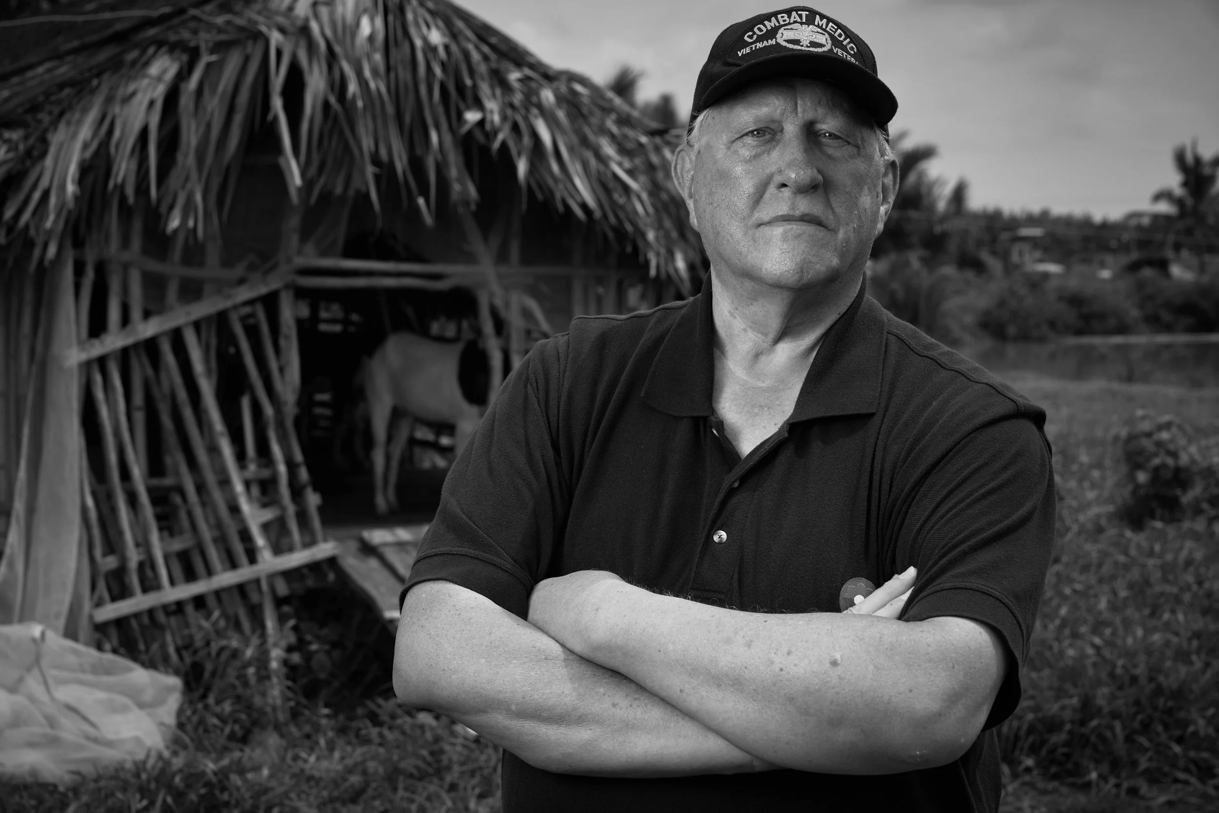 Black and white photograph of a man outdoors with crossed arms, wearing a cap that says 'Combat Medic Vietnam Veteran', with a thatched roof structure and trees in the background.
