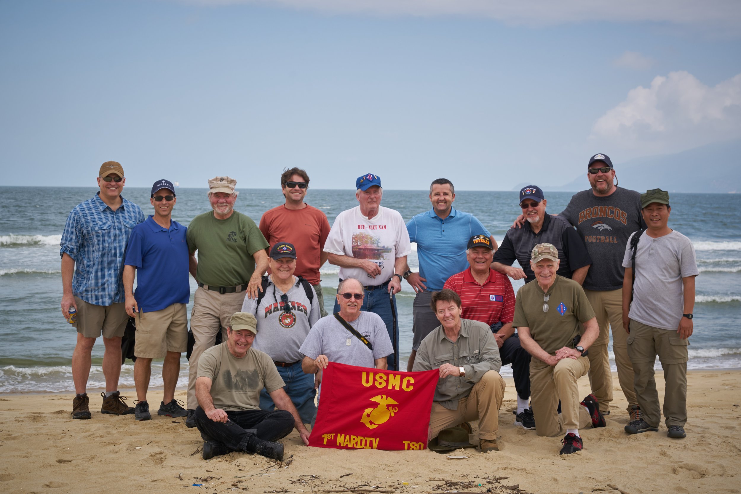 Group of men on a beach holding a USMC banner, with the ocean in the background.