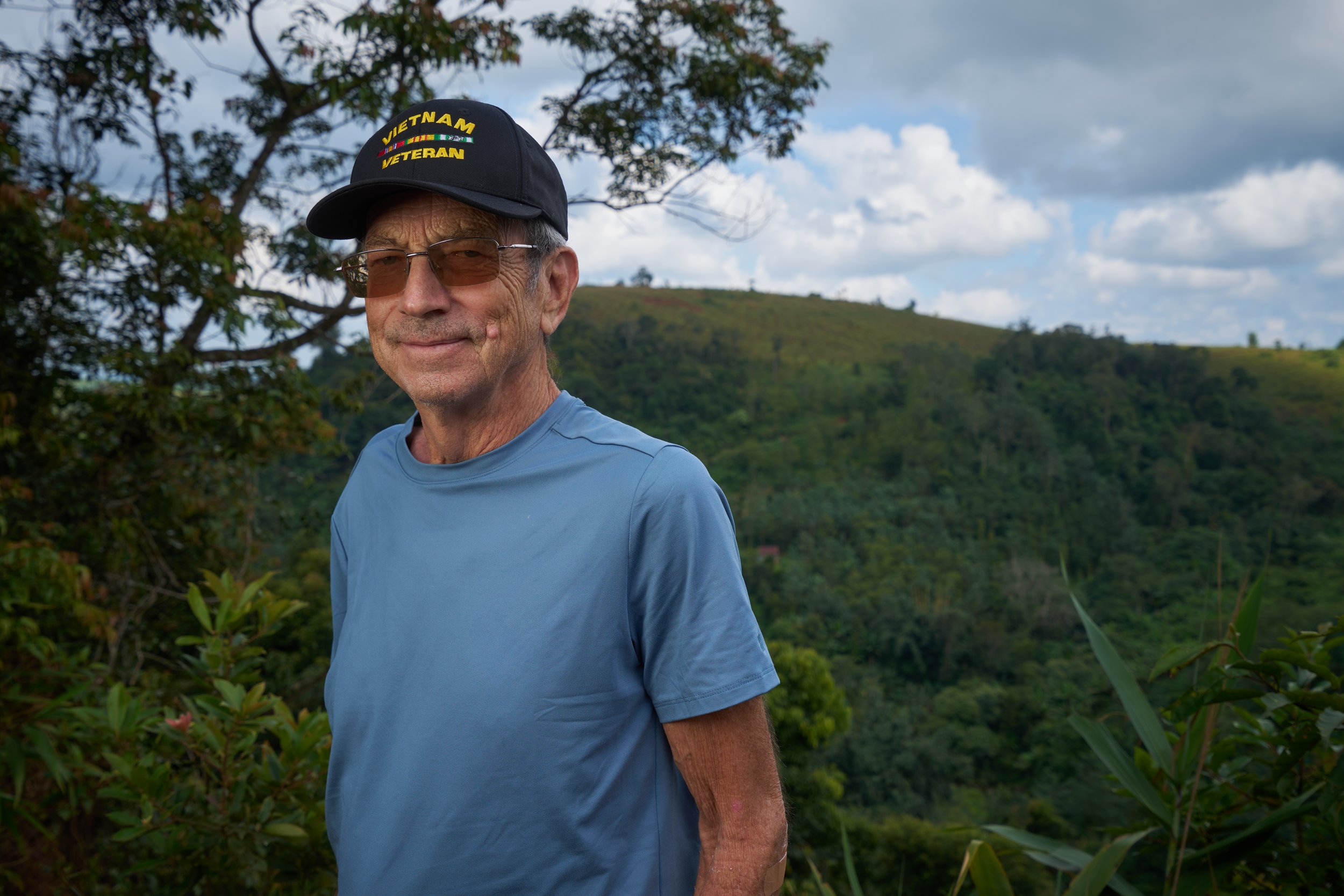 Man wearing a Vietnam veteran cap and glasses, standing outdoors in a lush green landscape.