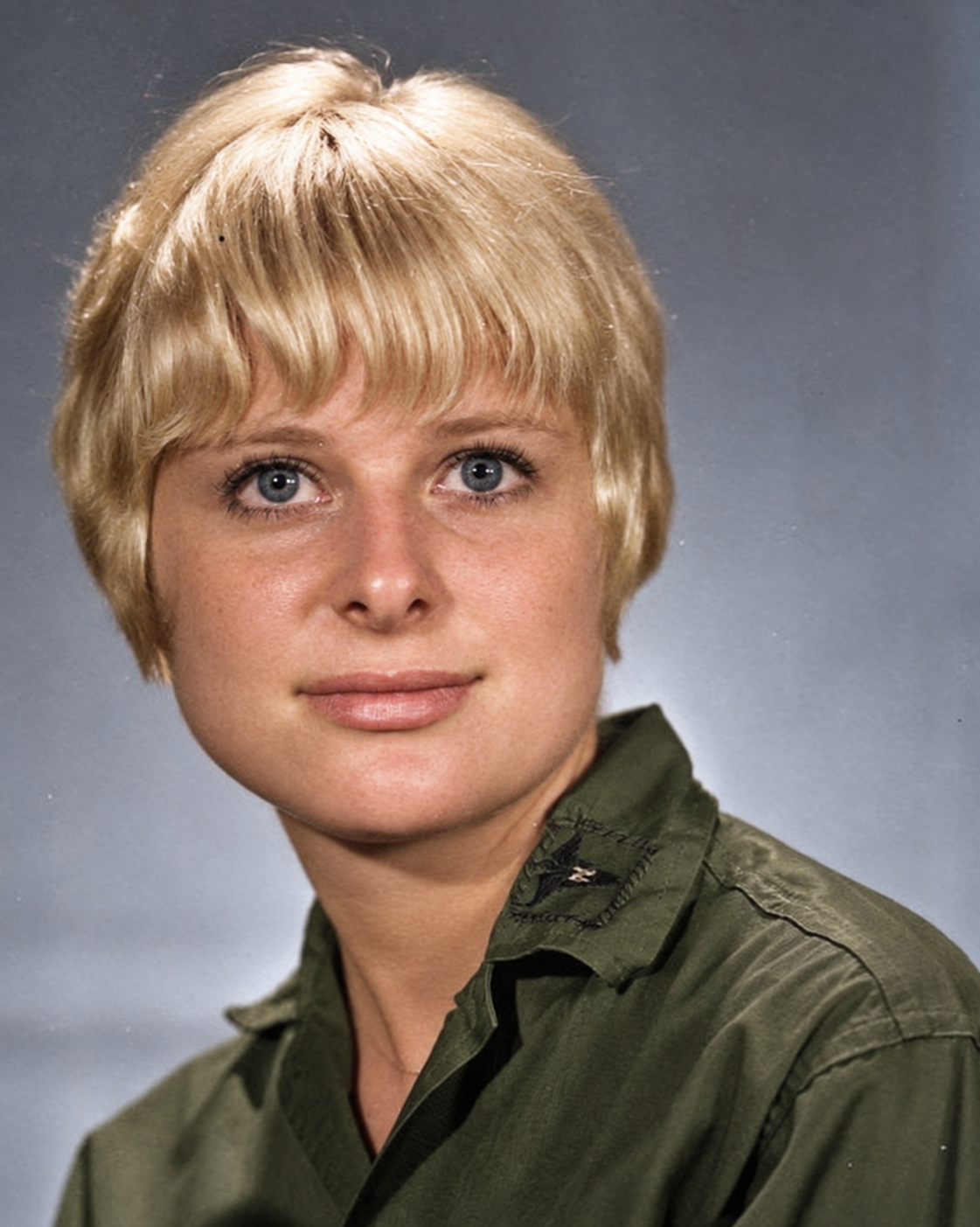Portrait of a woman with short blond hair, blue eyes, and wearing an olive green shirt against a neutral background.