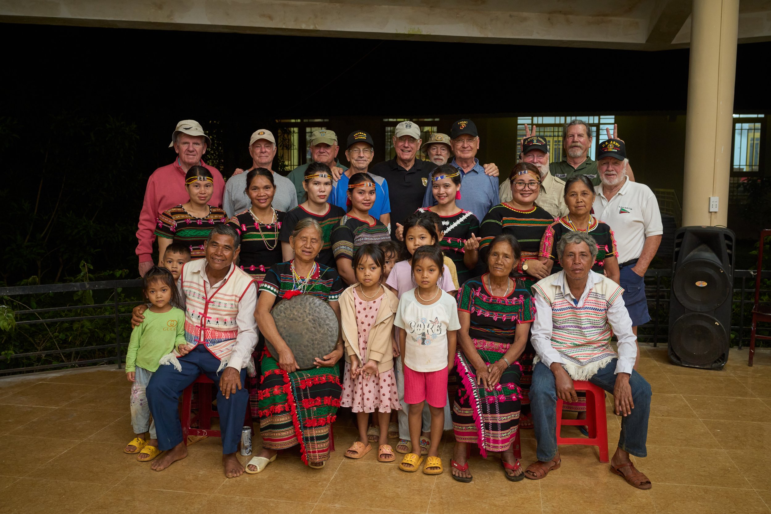 Group photo of diverse people, including children and adults, some wearing traditional clothing, posing indoors with tiled floor and dark background.