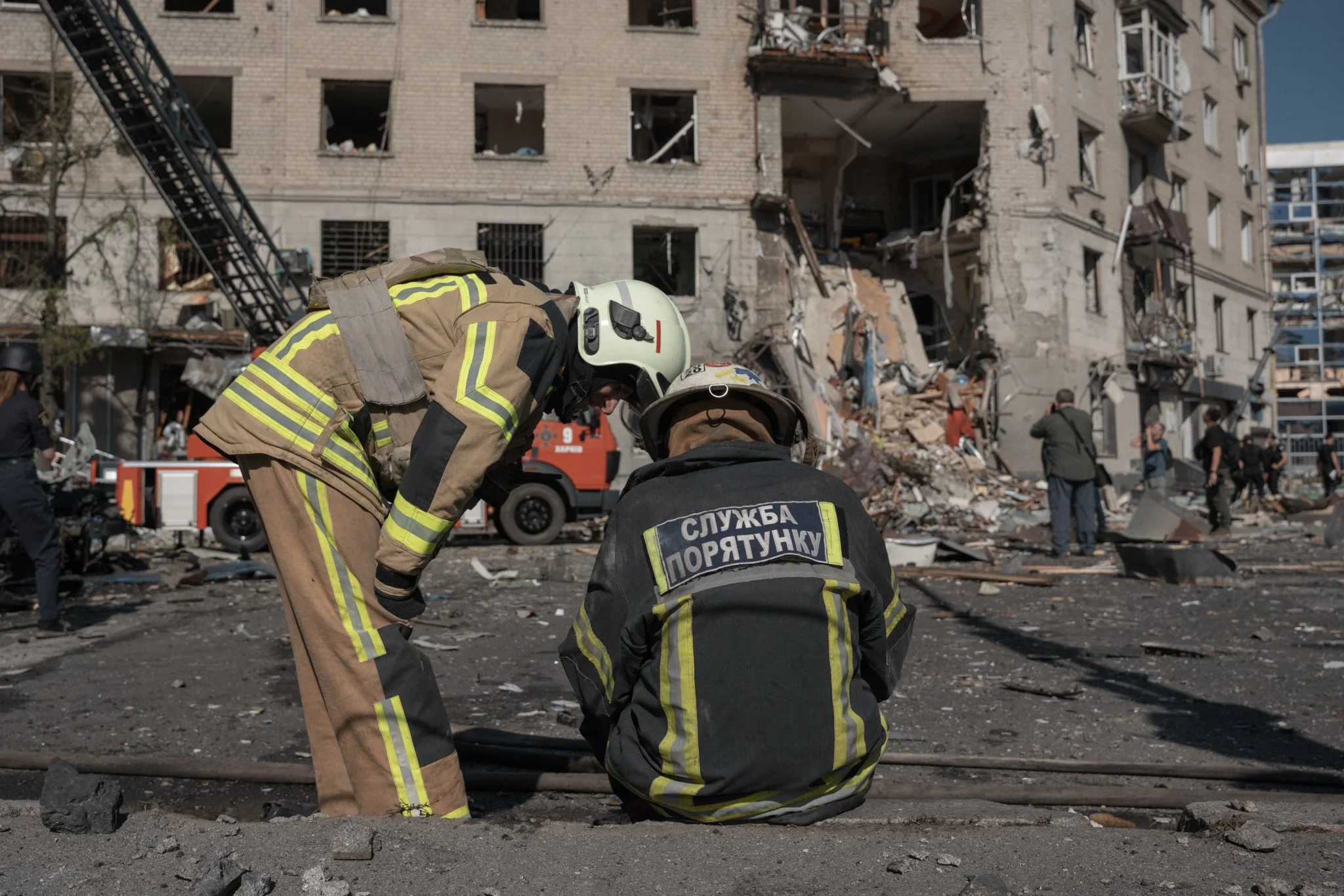 Firefighters in protective gear working in front of a damaged multi-story building with broken windows and debris, suggesting a recent disaster or rescue operation.