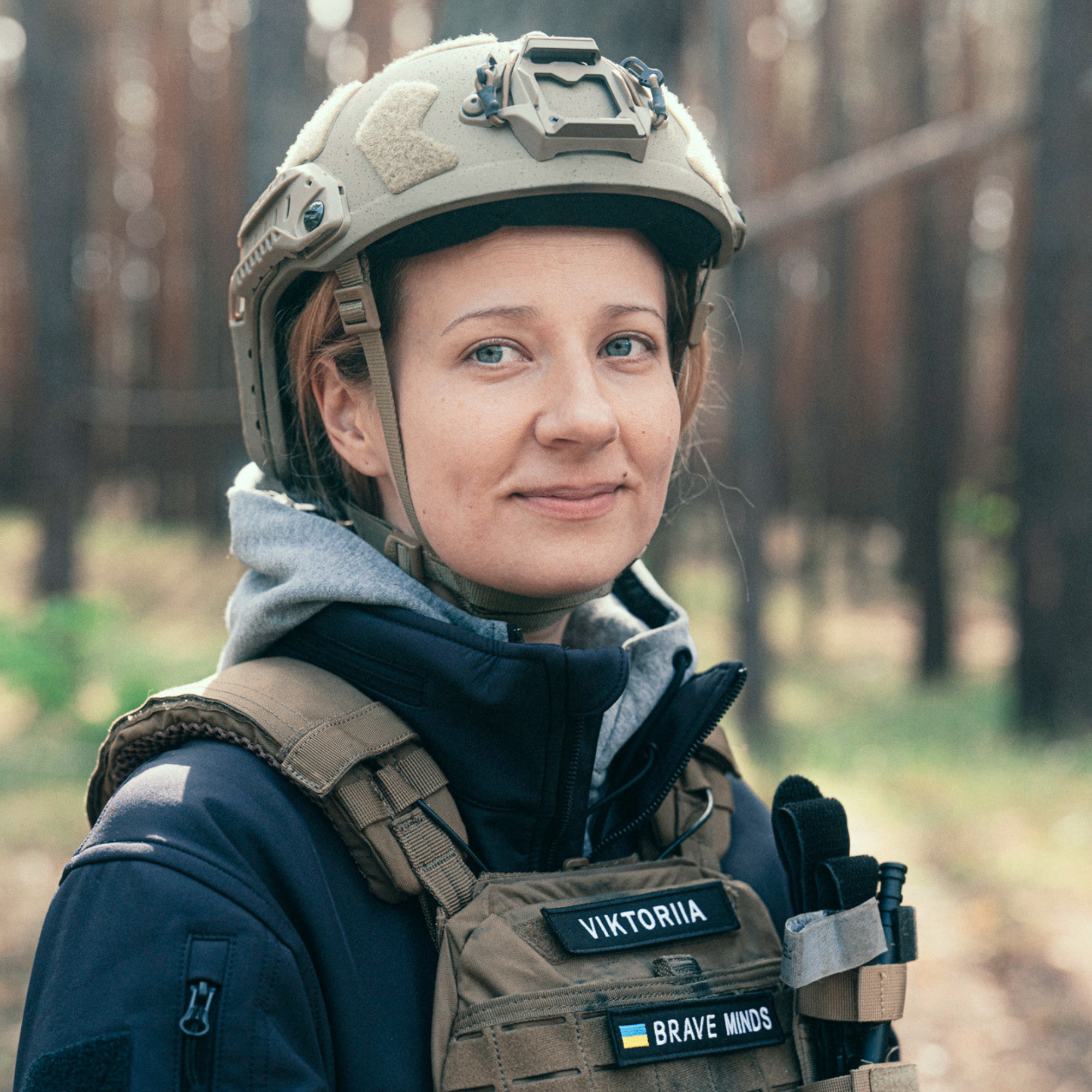 A woman in military gear wearing a tan tactical vest and a helmet outdoors with a wooded background. The vest has patches with the name Viktorija, Brave Minds, and a Ukrainian flag.