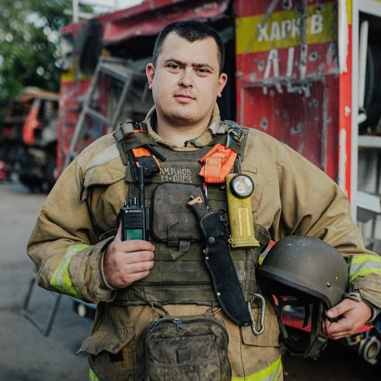 Male firefighter in uniform holding a helmet and a radio, standing in front of a fire truck.