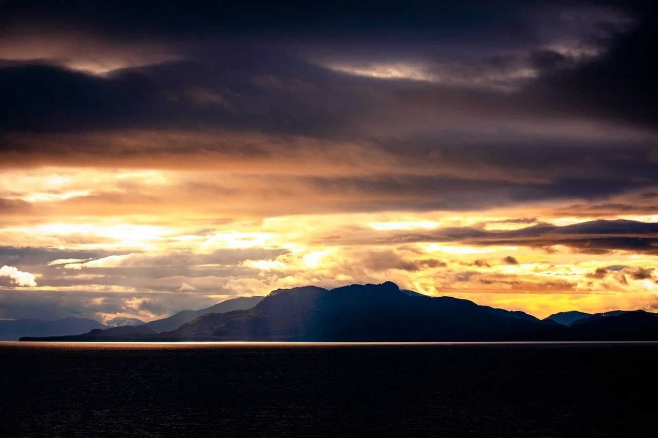 Glimmer of Hope.
.
As our ship glided through the Inside Passage, I woke before dawn to a world shrouded in darkness and clouds. Then, without warning, a thin ribbon of sunlight appeared along the horizon, the only break in the encompassing gloom. As