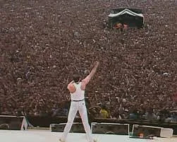 A defining Live Aid moment captures a rock frontman in white standing before a sea of humanity, arm lifted as tens of thousands respond in unison during a historic concert.