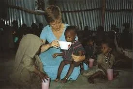 A humanitarian aid worker sits inside a modest shelter, feeding a small child while other children sit close by with cups, illustrating hands-on relief work and human connection during a humanitarian emergency.