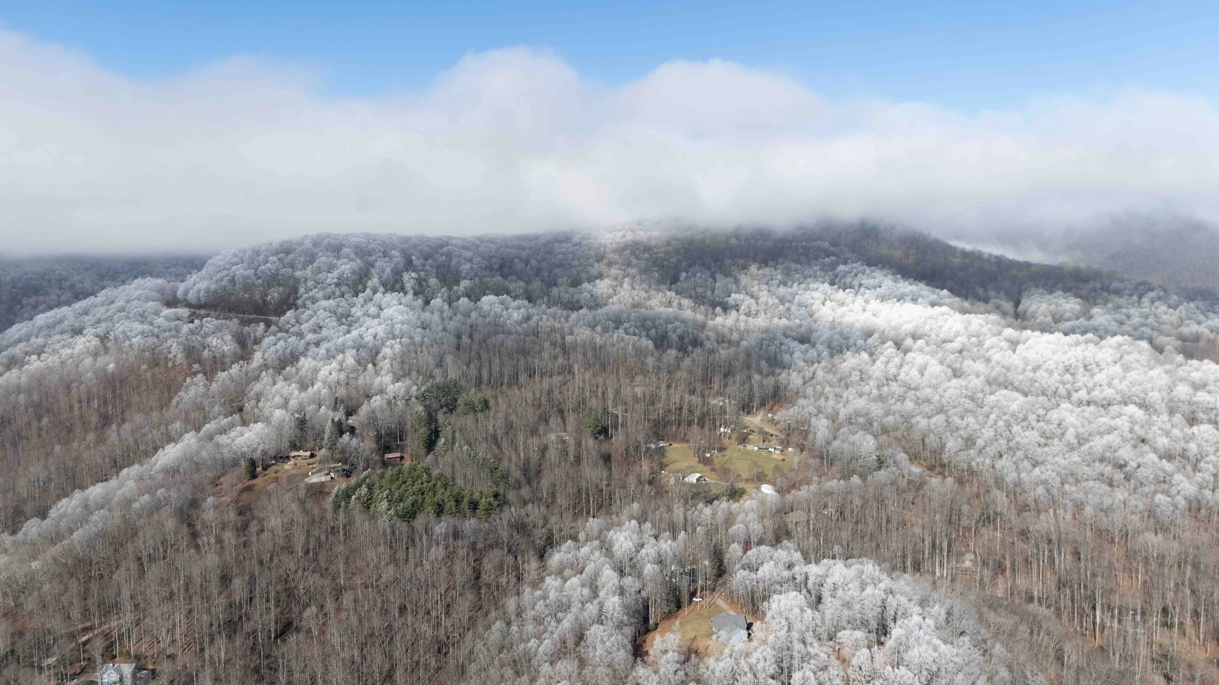 Aerial view of a snow-covered mountain landscape with a small rural village and trees in winter, with a partly cloudy sky and low-hanging clouds near the mountain peaks.