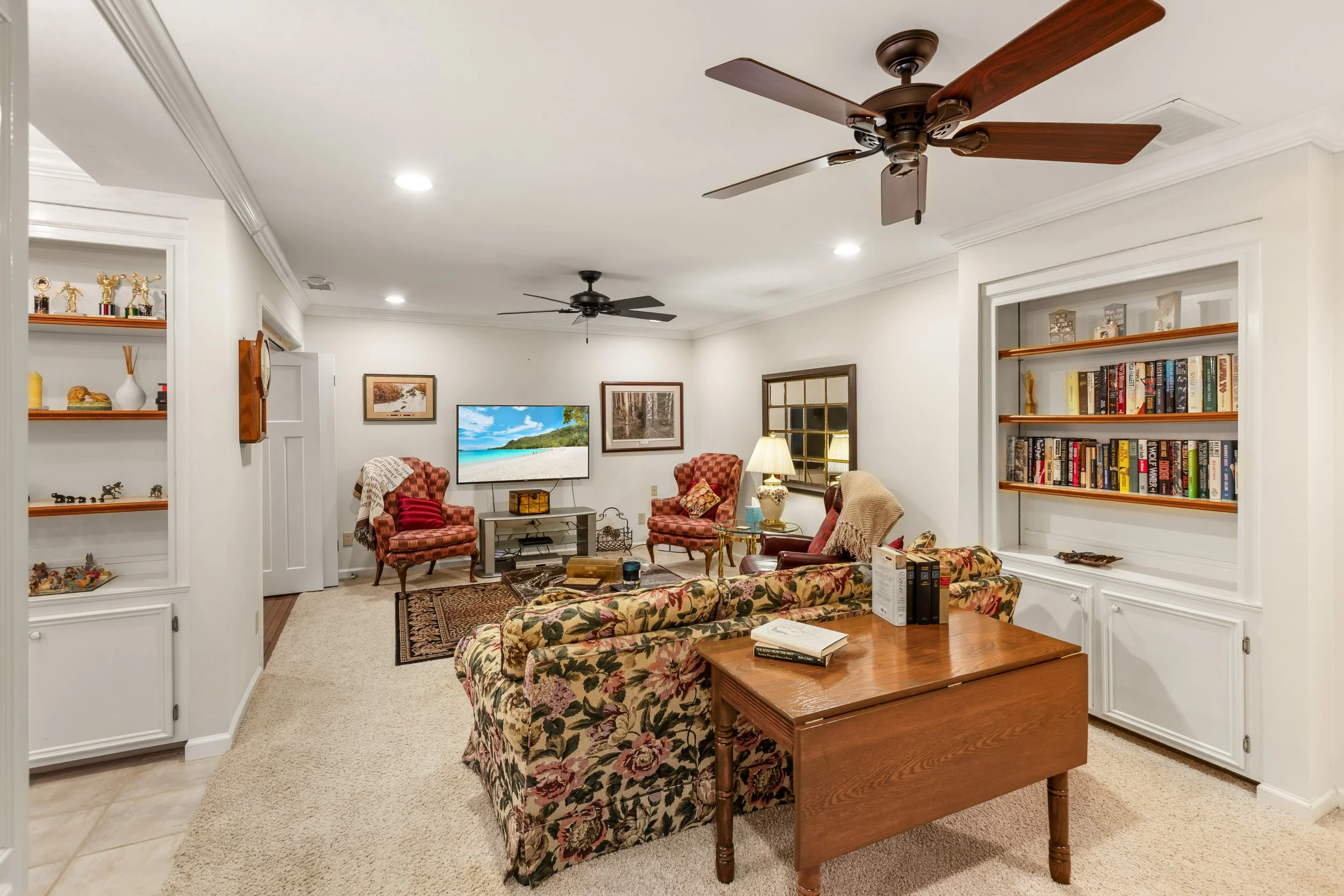 Living room with a floral sofa, wooden side table, armchairs, a TV, and built-in bookshelves