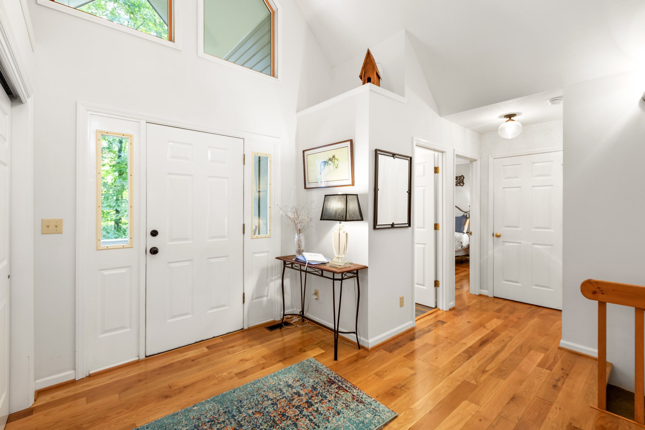 Entryway with white door, small table with lamp and decorative items, framed art, and hardwood floors. A window near the ceiling and views of greenery outside.