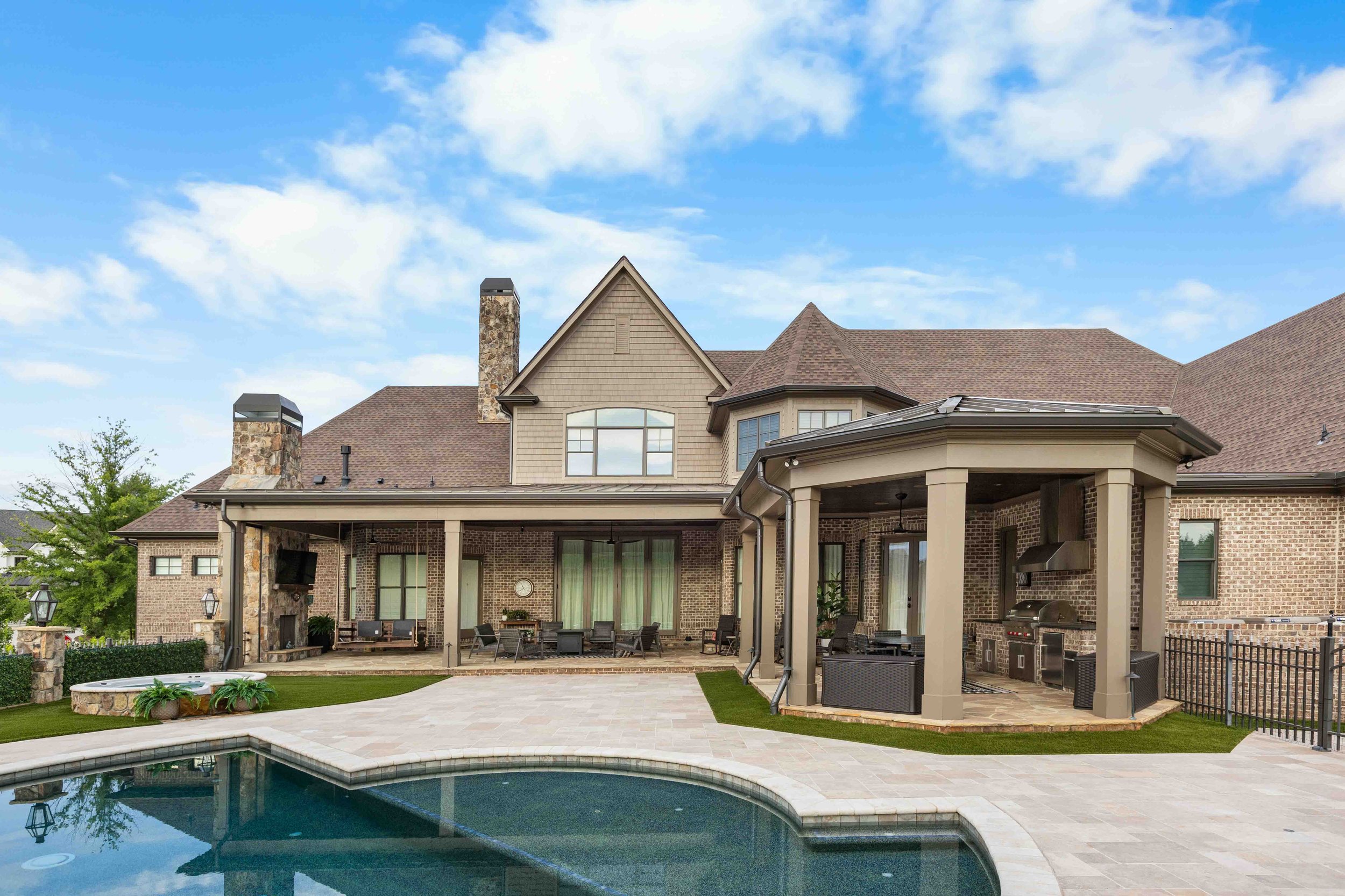 Backyard with swimming pool, outdoor seating, outdoor kitchen, and patio, with a large brick house and blue sky in the background.