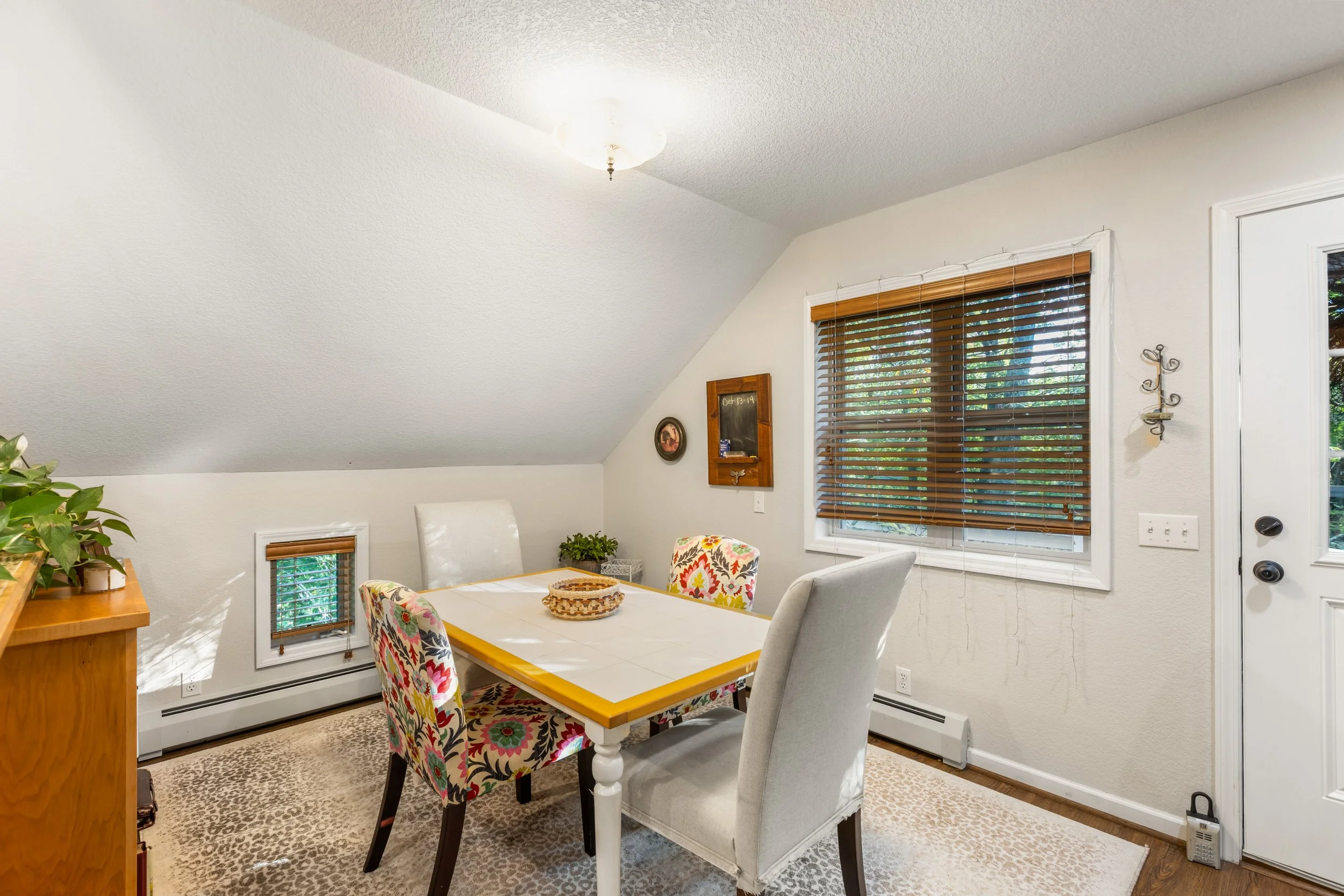 Dining room with a rectangular table, four chairs (two floral and two plain), a window with blinds, a wall decor, and a small plant on the table. The room has a sloped ceiling, a ceiling light, and a small window near the floor.