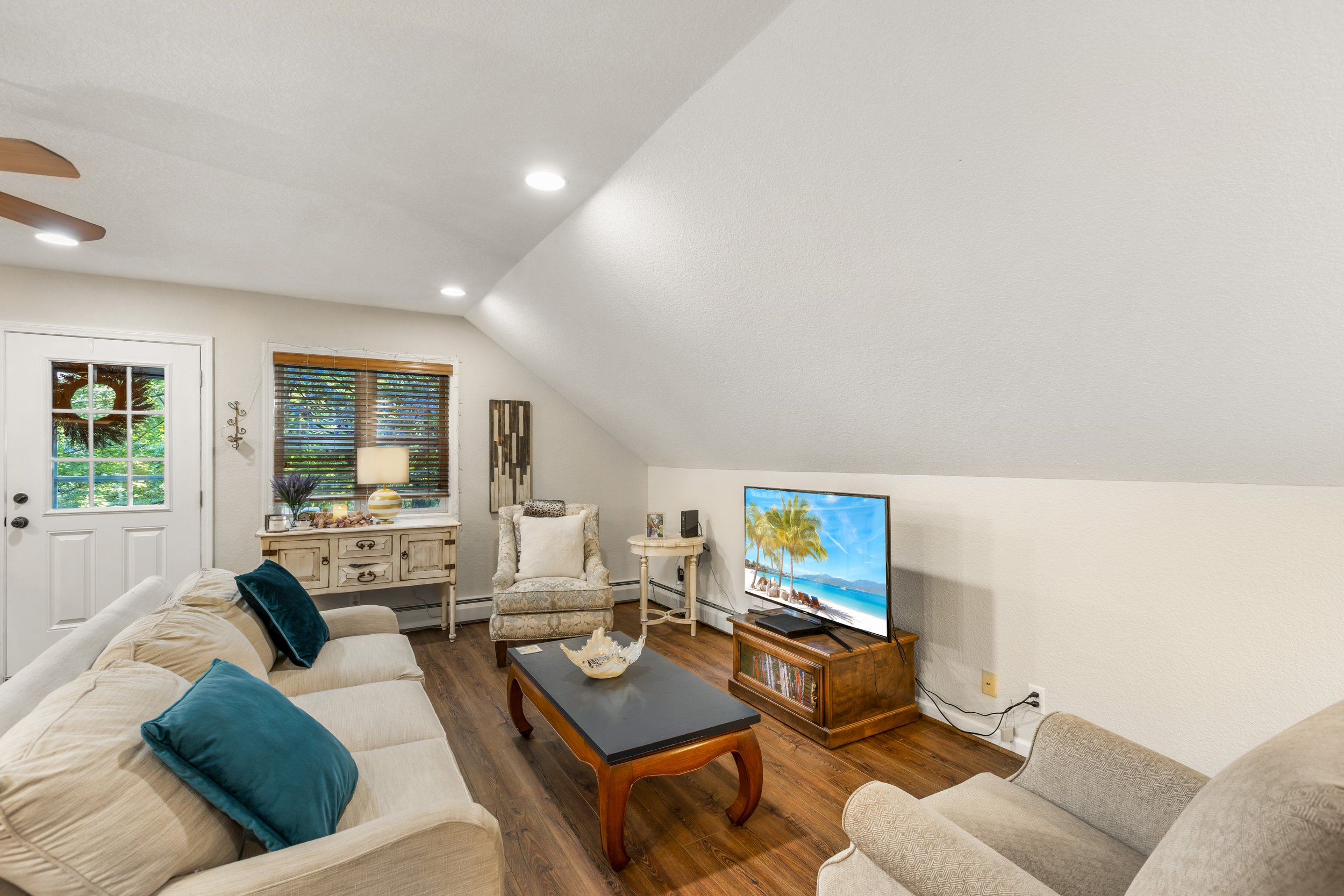 Living room with beige sofa, armchair, coffee table, TV on wooden stand, decorative items, window with blinds, door with window, wood flooring, ceiling with recessed lighting.