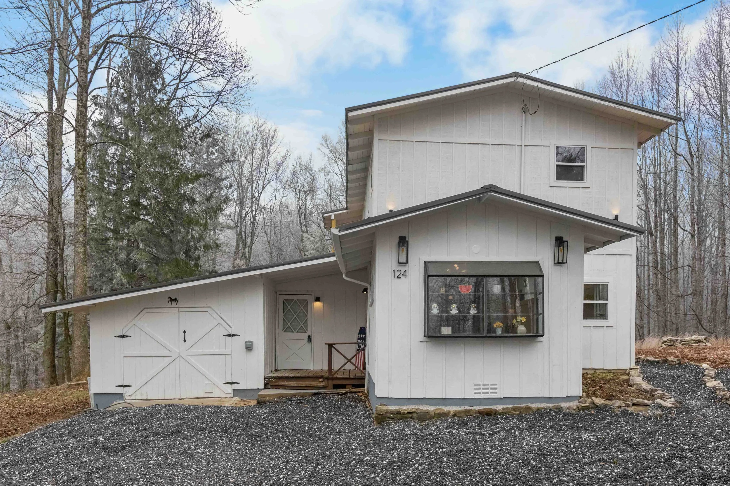 A white two-story house with a garage, wooden deck, and front porch, surrounded by trees and a gravel driveway.