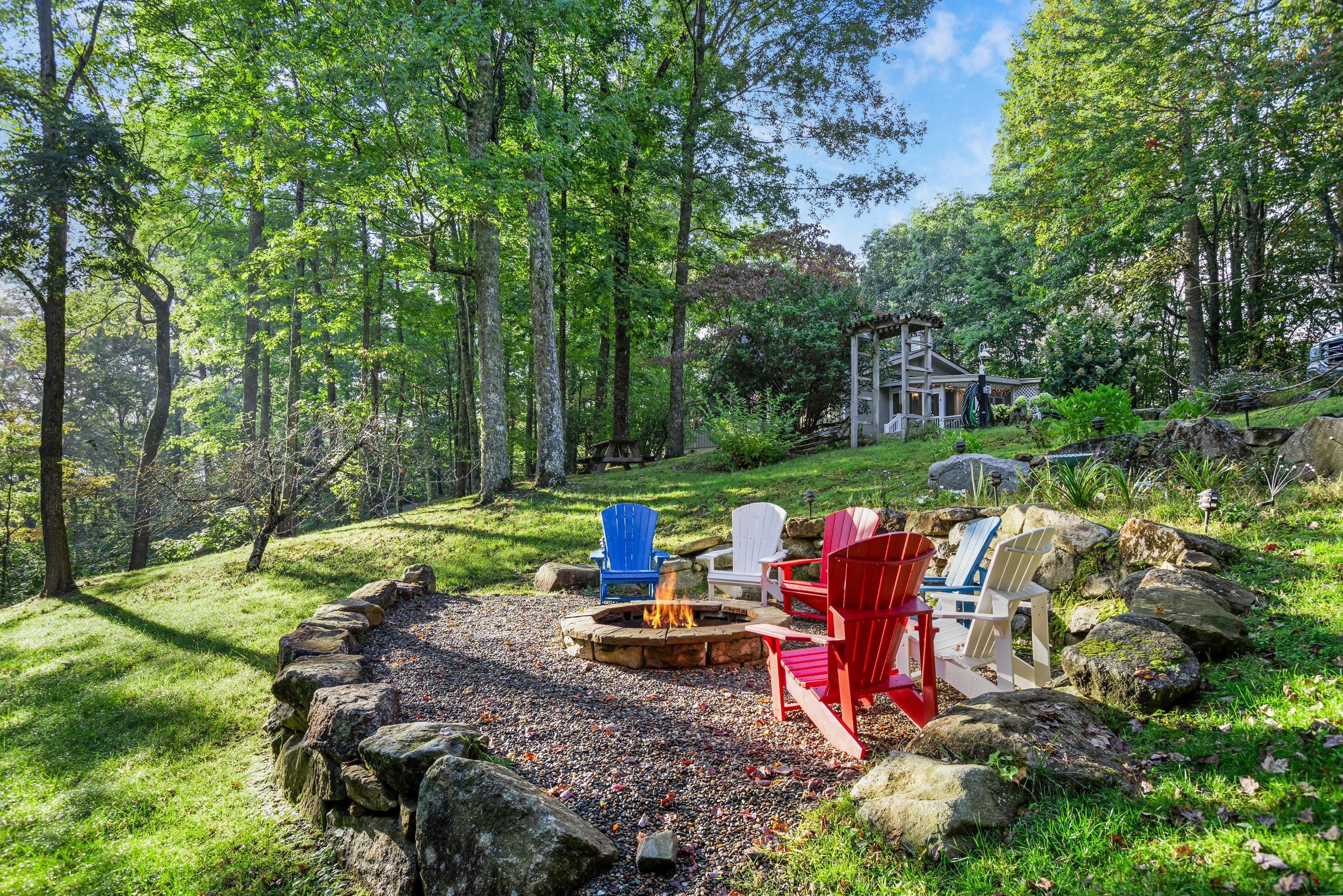Colorful Adirondack chairs arranged in a circle around a small fire pit in a wooded backyard with lush green grass and trees.