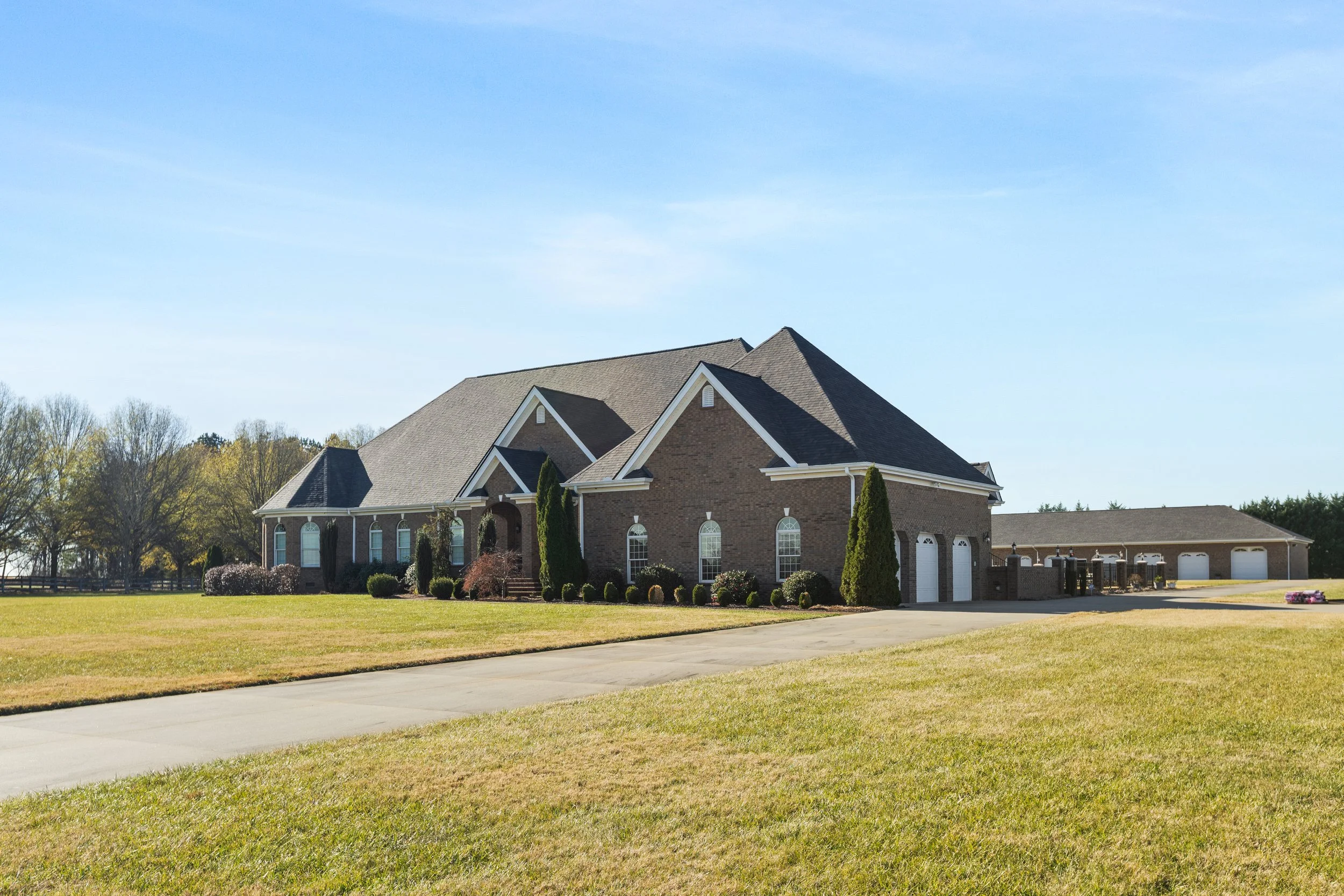 Large brick house with multiple gabled roofs, surrounded by a well-maintained lawn, trees, and driveway, under a clear blue sky.