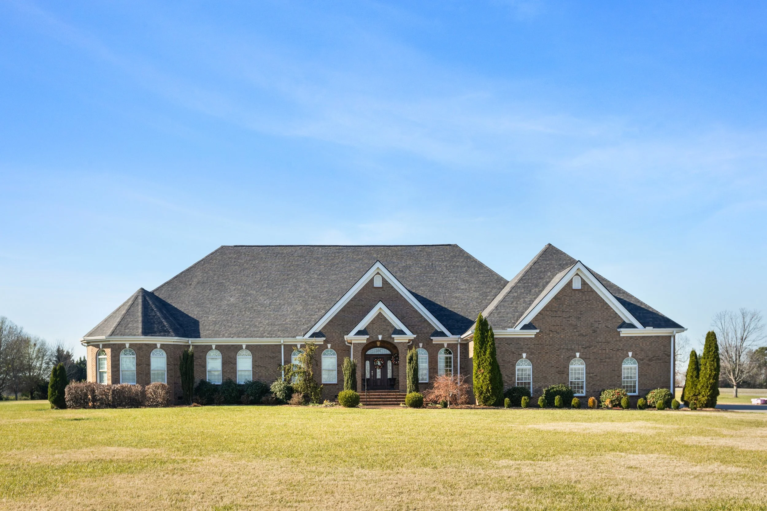 A large brick house with white-trimmed arched windows, a peaked roof, and a front entrance with steps and tall trees and bushes in front, set on a spacious grassy lawn with a clear blue sky in the background.