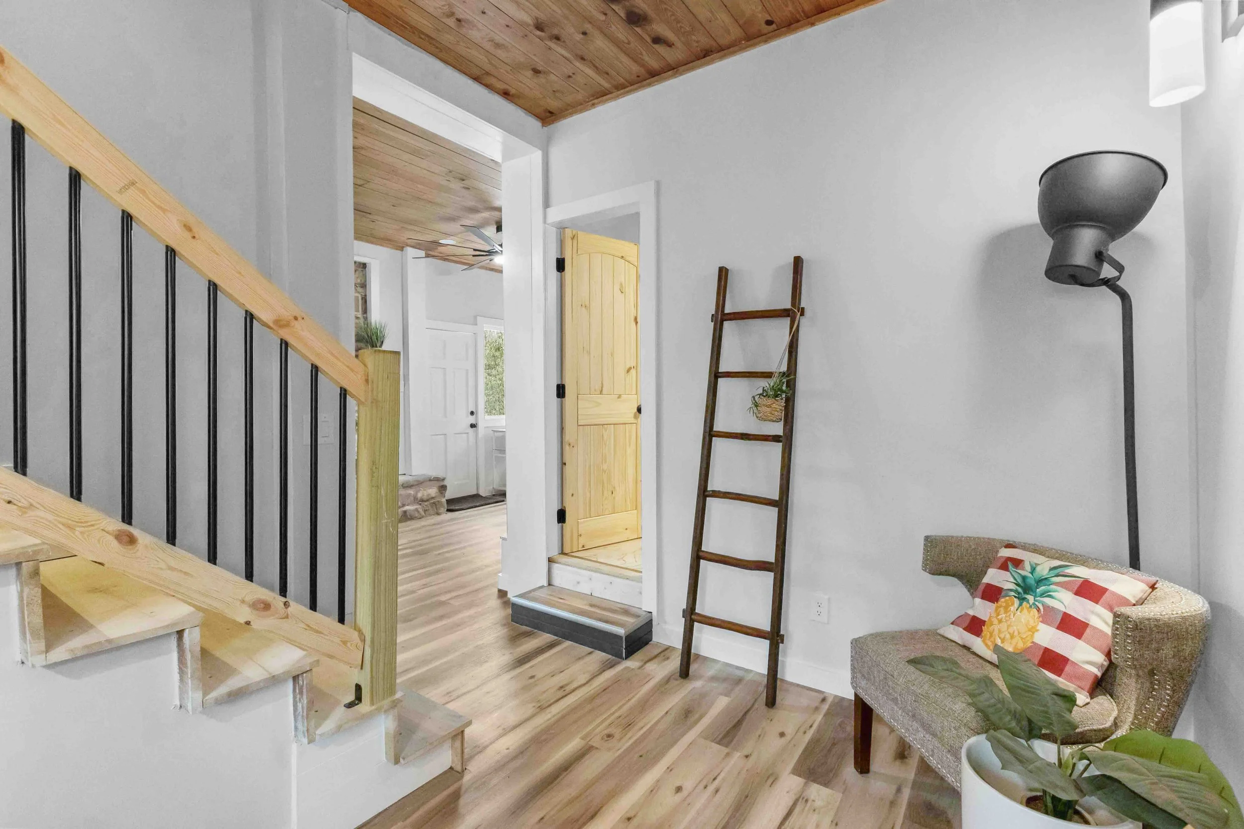 Interior view of a home with wooden floors, white walls, a staircase with a wooden railing, a doorway with a wood door, a ladder leaning against the wall with a small plant hanging from it, a cushioned chair with tropical pillow, a potted plant, and 
