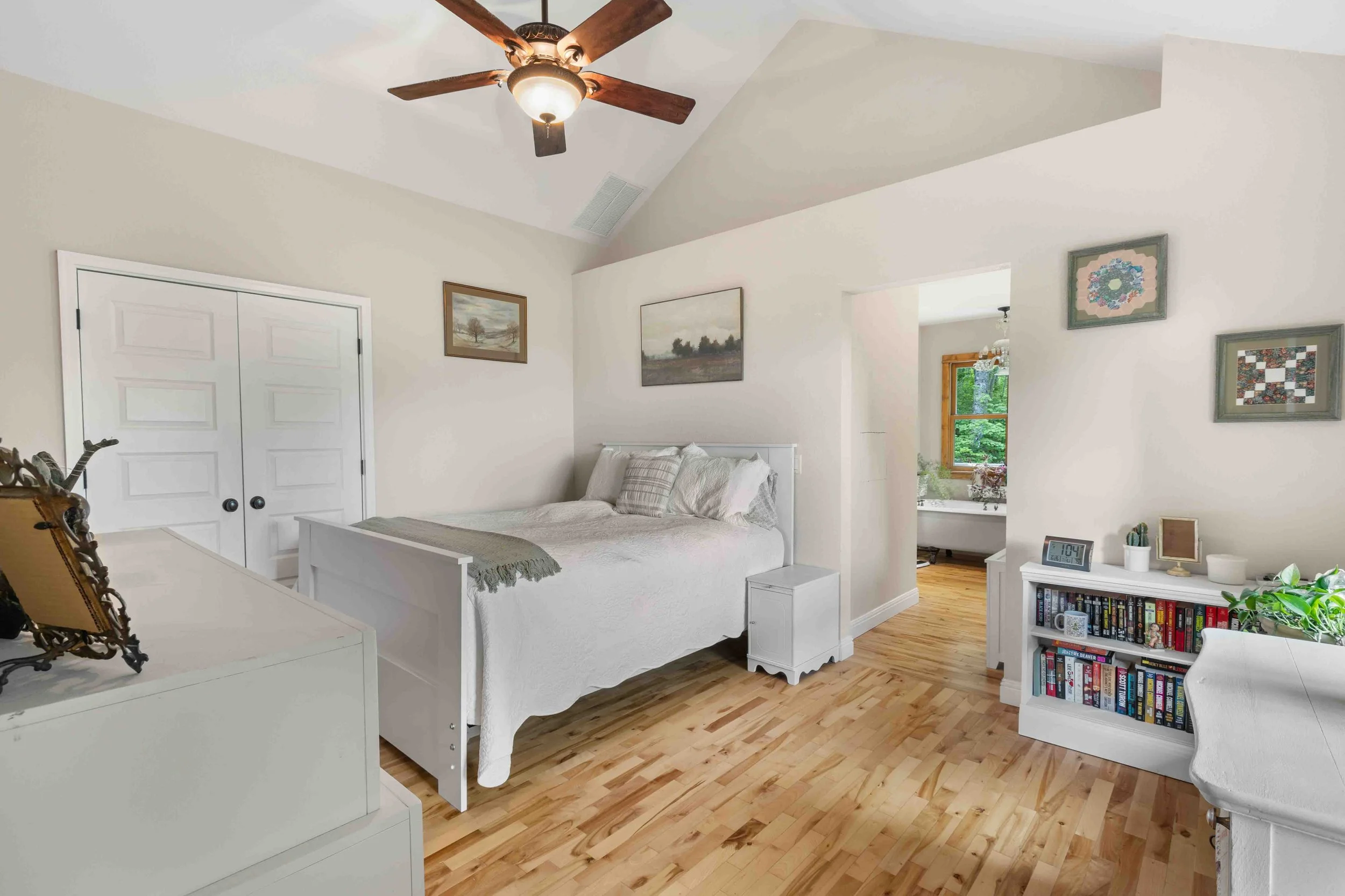 A bedroom with a white bed, wooden hall, and various paintings and pictures on the walls. There is a ceiling fan and an open doorway leading to a bathroom with a bathtub and window.