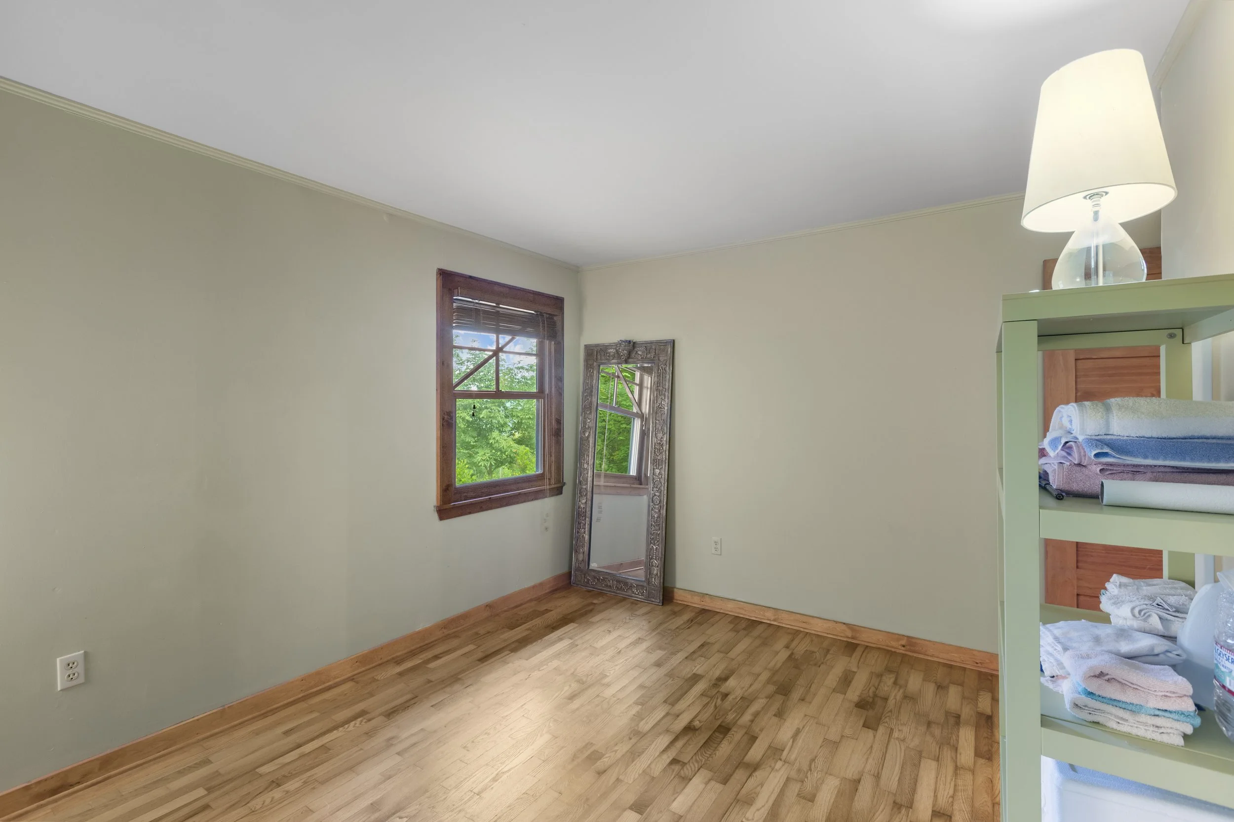 Empty room with wooden floor, two windows, a large mirror leaning against the wall, and a small shelf with folded towels and a lamp.