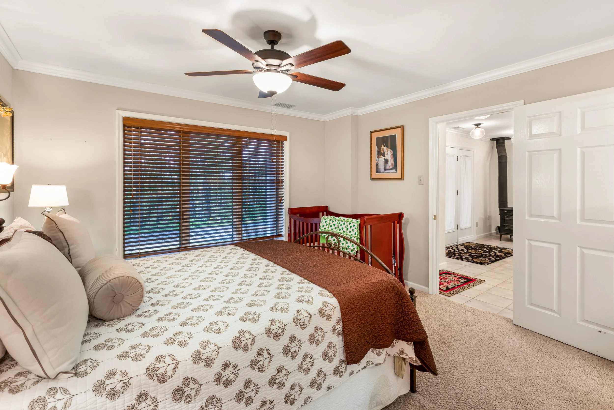Bedroom with a bed, window with wooden blinds, ceiling fan, and an open doorway leading to an entryway with tiled flooring and a stove.