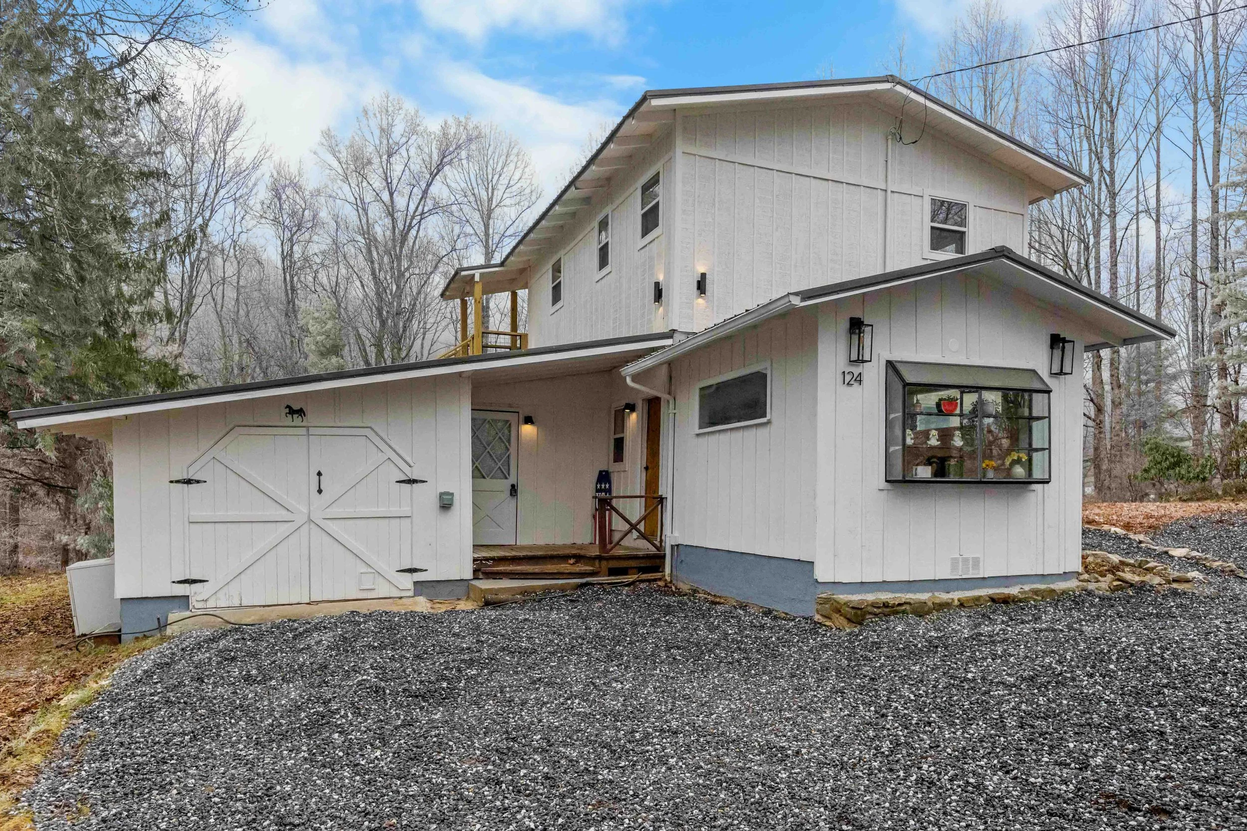 A modern two-story white house with a gravel driveway, surrounded by leafless trees, featuring several windows, a garage with a barn-style door, and a small porch with a wooden railing.